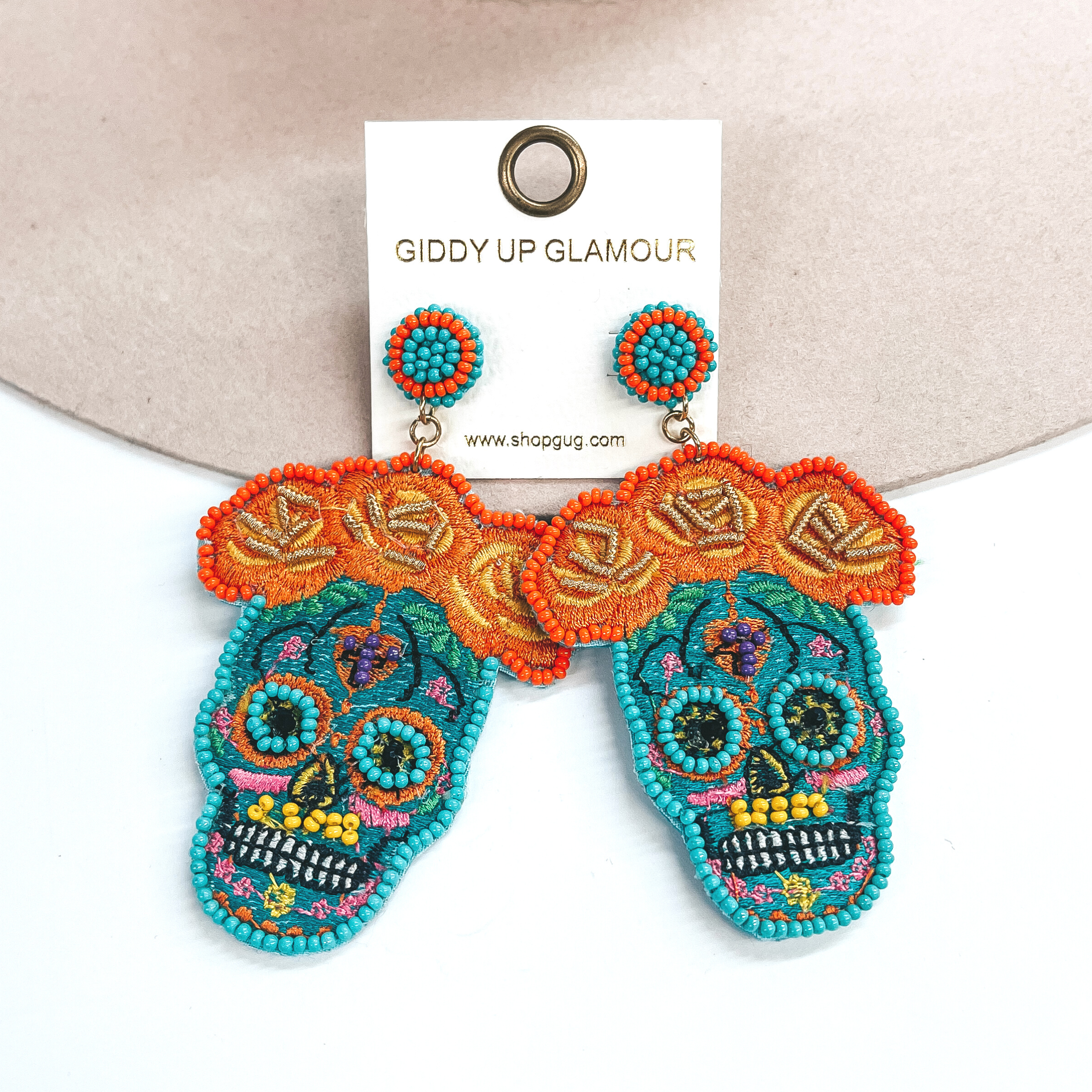 These are sugar skull post earrings in turquoise and other colors. There are orange  flowers in the top with beads and stitching. The skull part has turquoise beads  around the eyes, yellow beads in the mouth, and black beads in the eyes.  The rest has multicolored stitching all around. These earrings are taken on  a beige hat brim and on a white background.