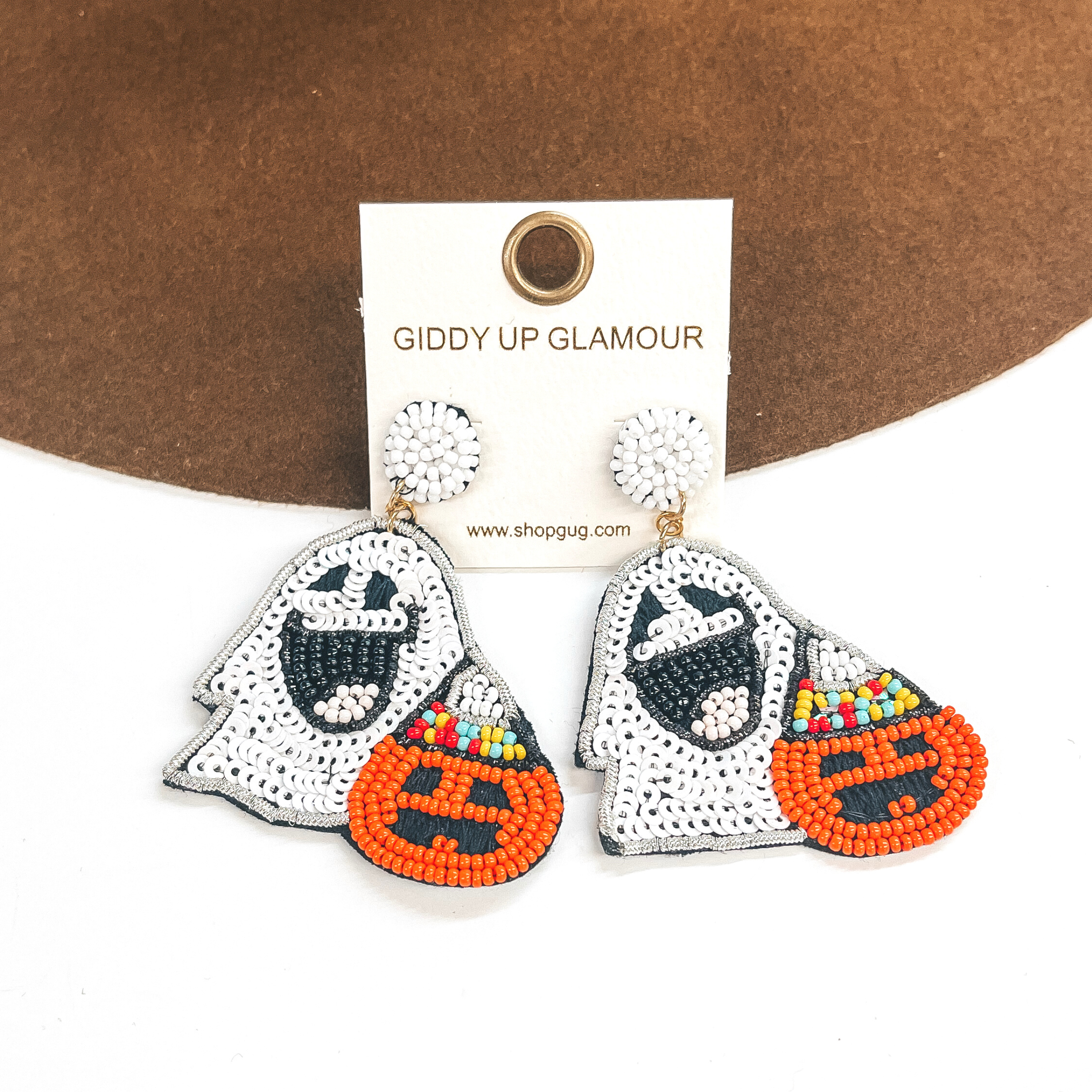 Pictured on a white and brown background are beaded white ghost earrings. These earrings also include a orange pumpkin that has a party hat on. 