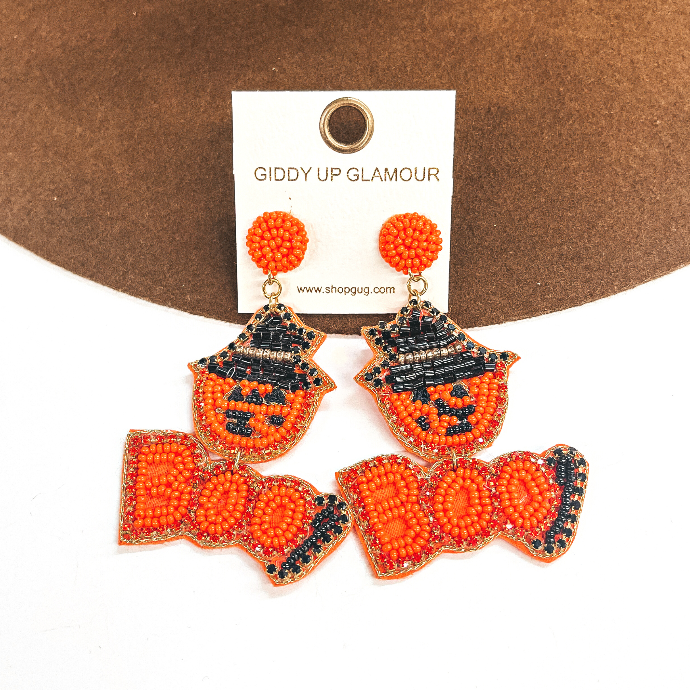 Pictured on a white and brown background are beaded earrings with an orange pumpking with a black withc hat. This pumpkin also has an orange beaded pendant that spells out "BOO." 
