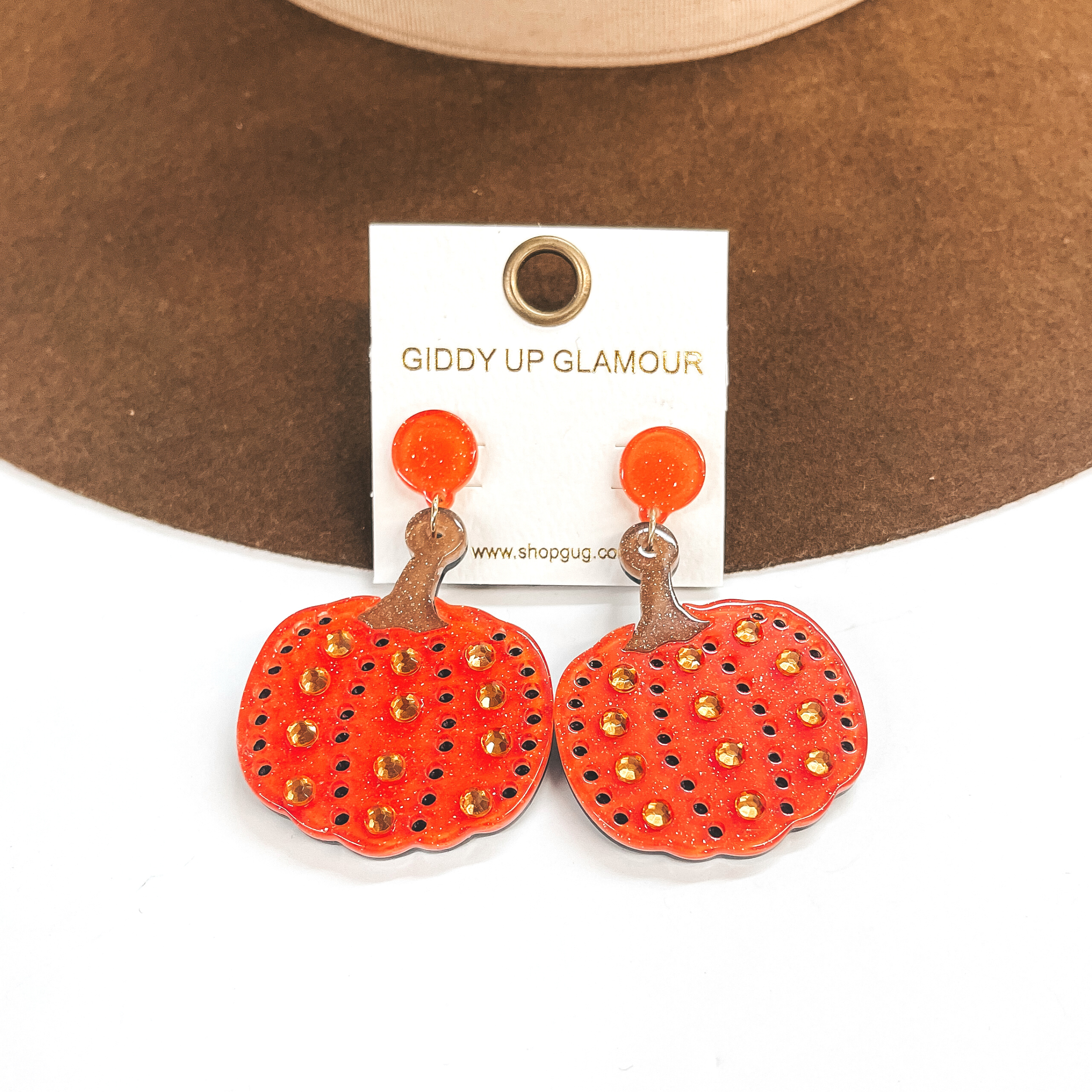 Pictured on a white and brown background are acrylic, orange glitter pumpkin earrings. These earrings include holes as and crystals as detailing.