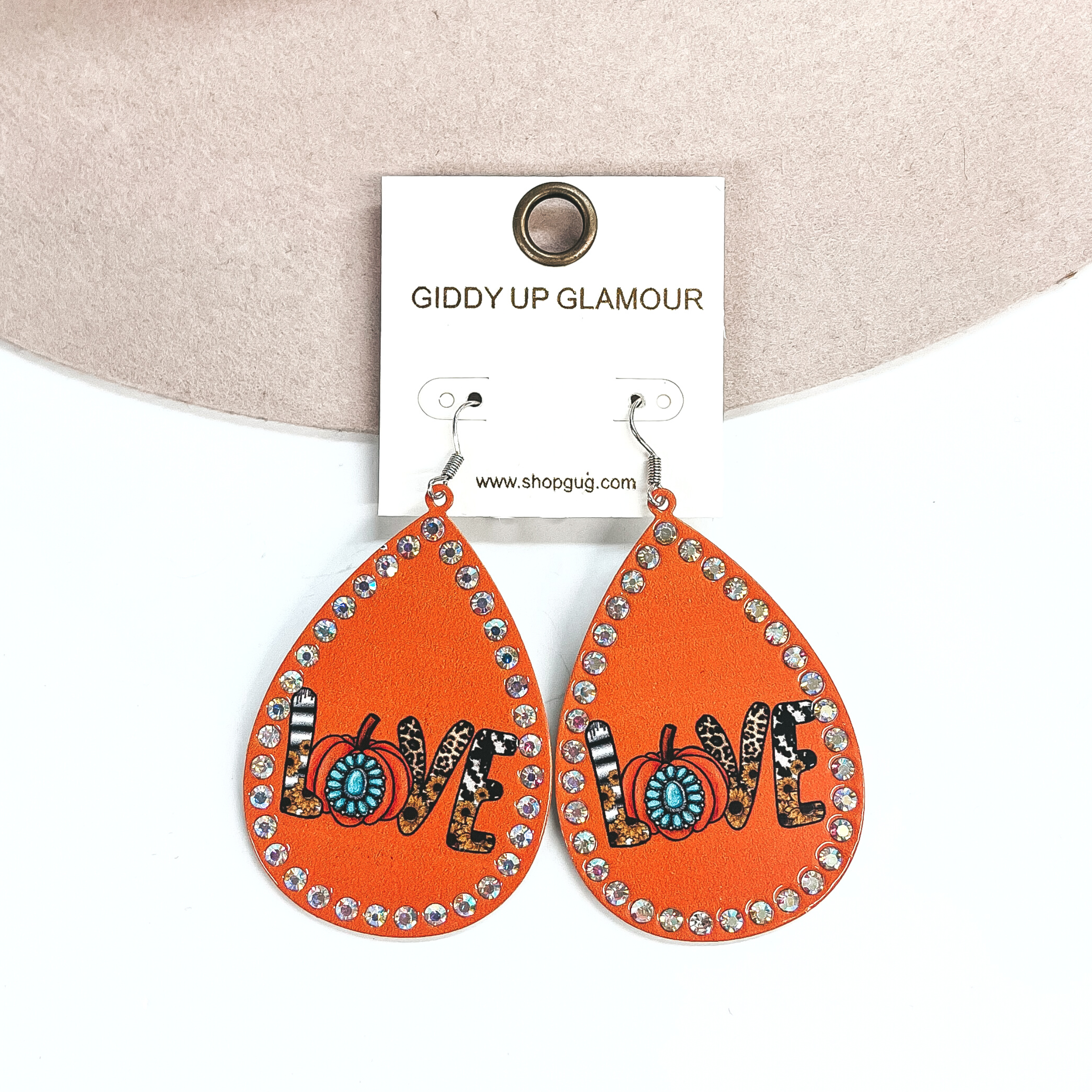These are teardrop earrings in orange with AB crystals all around. In the middle it says 'Love' , each letter has different prints and designs. The 'O' is an orange pumpkin with a turquoise concho in the center. These earrings are taken laying on a beige color hat brim and on a white background