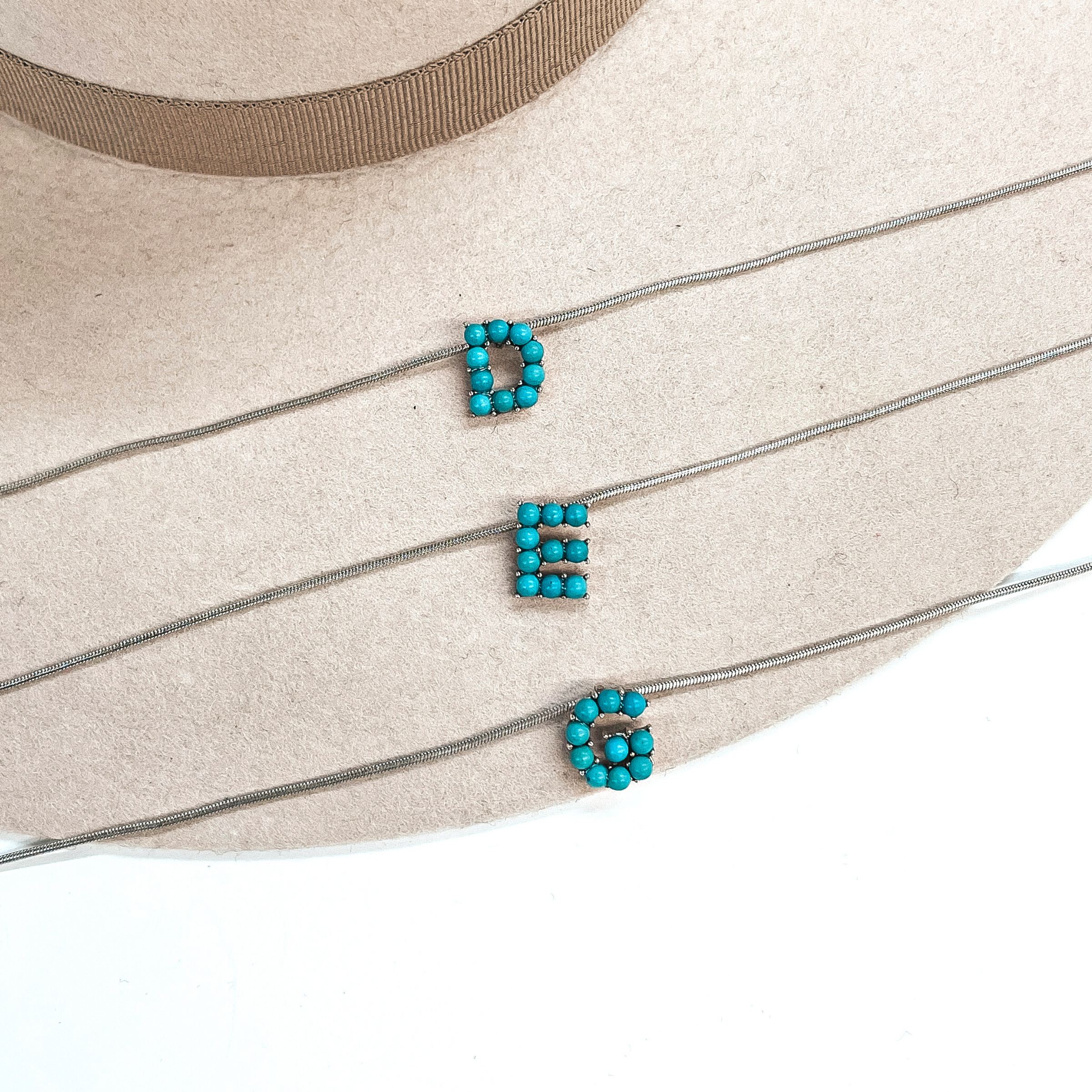 These are three turquoise stone initial necklaces with a thin, silver  snake chain. From top to bottom; D, E, G. These necklaces are taken  laying on a beige felt hat and on a white background.