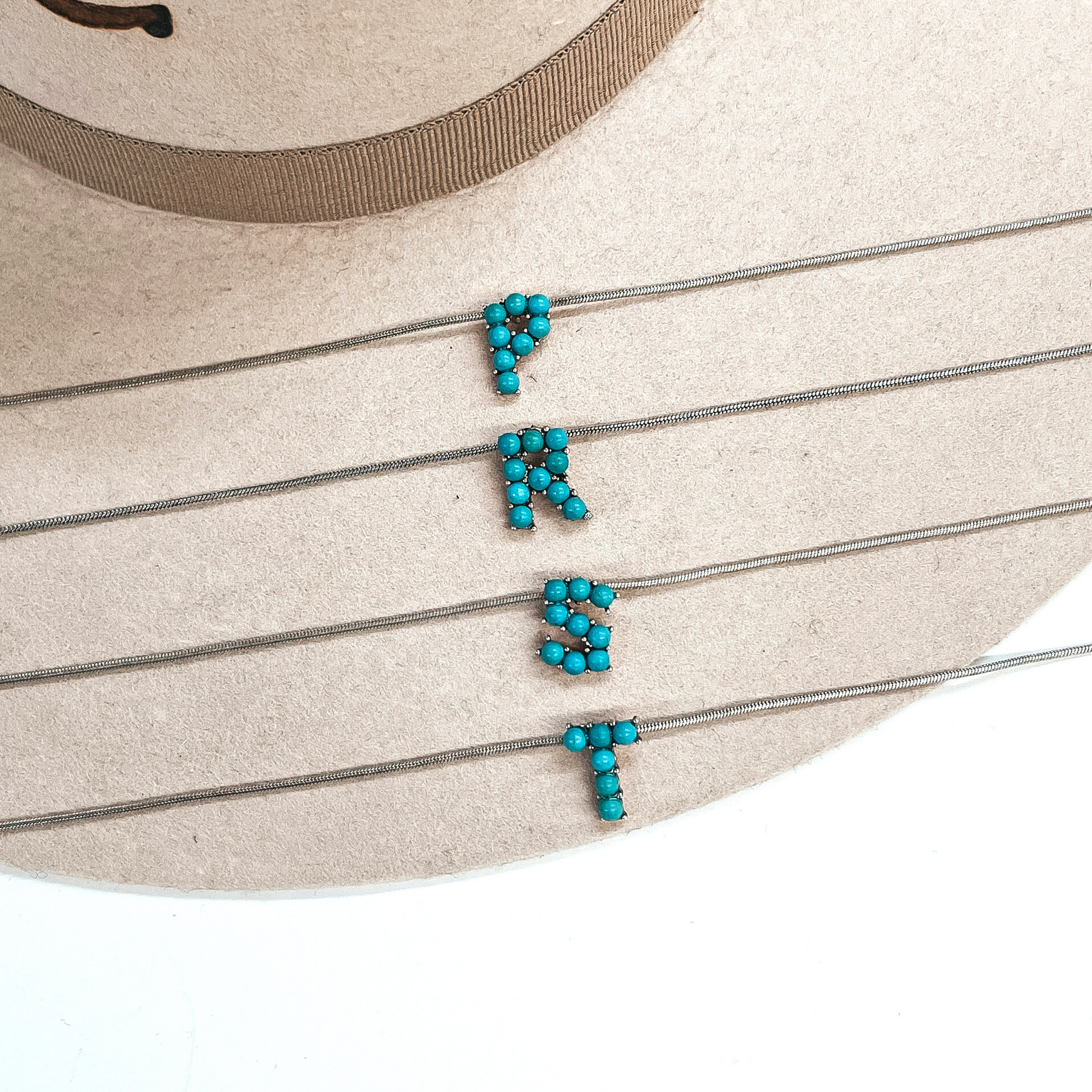 These are four turquoise stone initial necklaces with a thin, silver  snake chain. From top to bottom; P, R, S, T. These necklaces are taken  laying on a beige felt hat and on a white background.