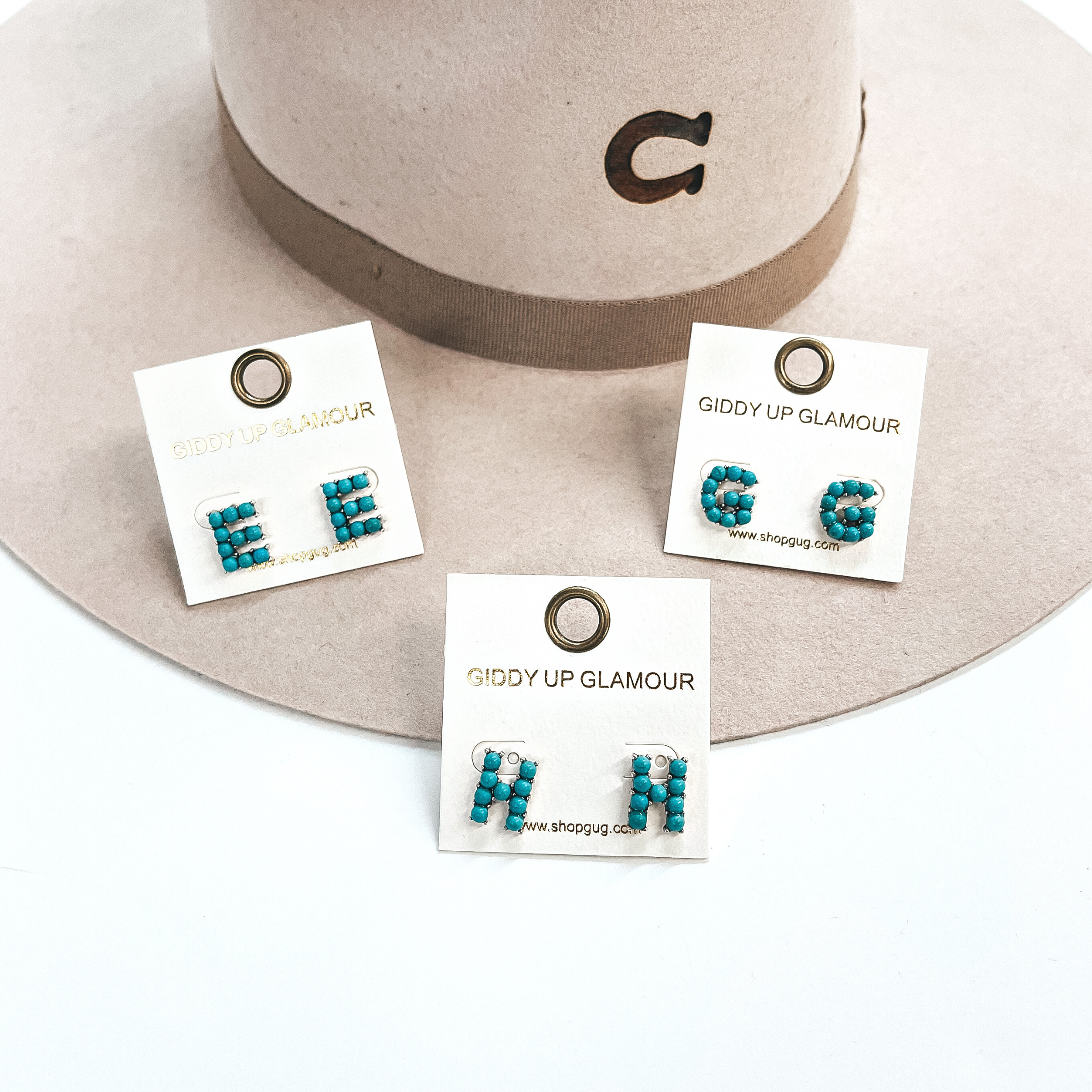 These are three pair of initial earrings with small turquoise stones in  a silver setting. From left to right; E,H,G. These earrings are placed  on a white Giddy Up Glamour card, they are laying on a beige, felt,  Charlie 1 Horse hat and on a white background.