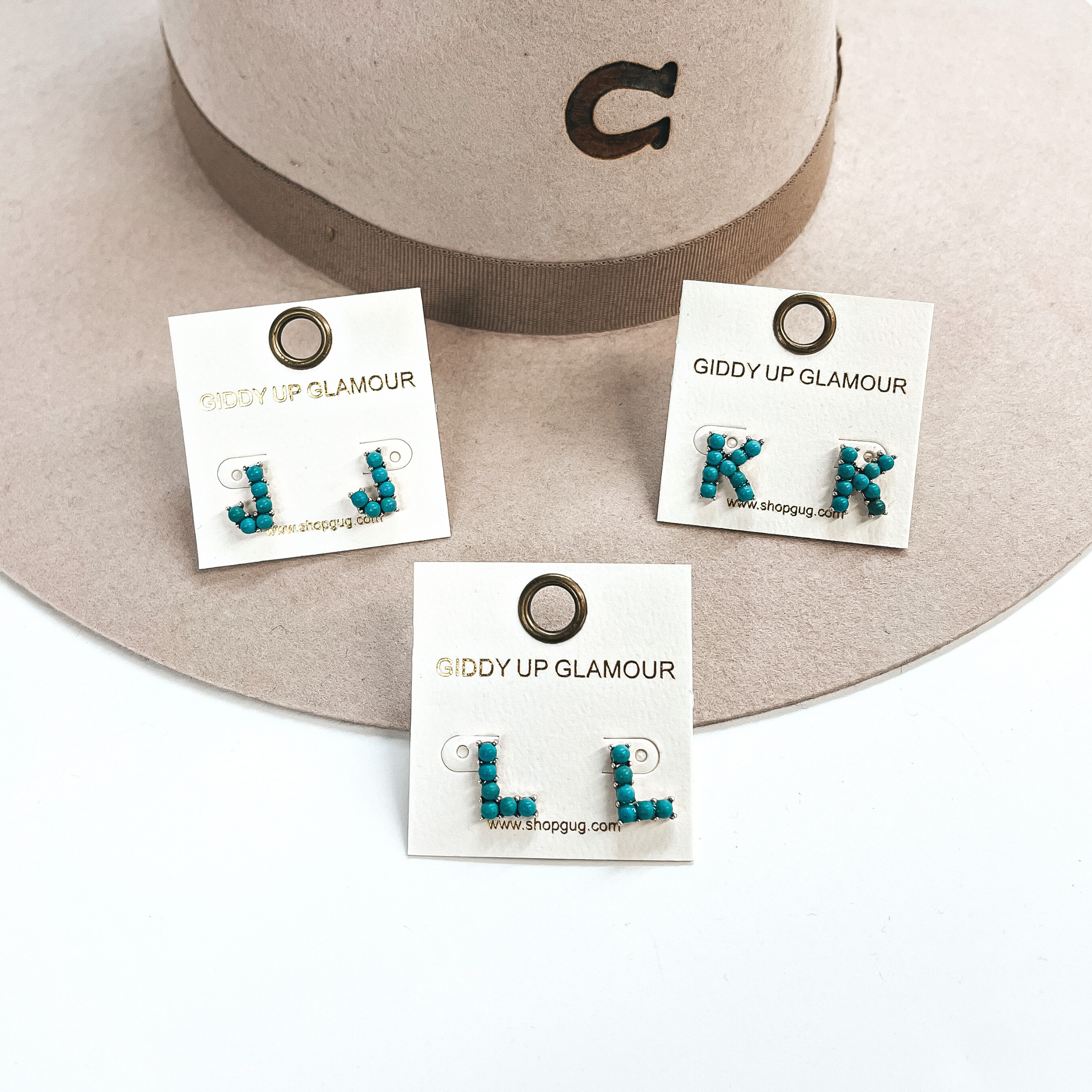 These are three pair of initial earrings with small turquoise stones in  a silver setting. From left to right; J,L,K. These earrings are placed  on a white Giddy Up Glamour card, they are laying on a beige, felt,  Charlie 1 Horse hat and on a white background.