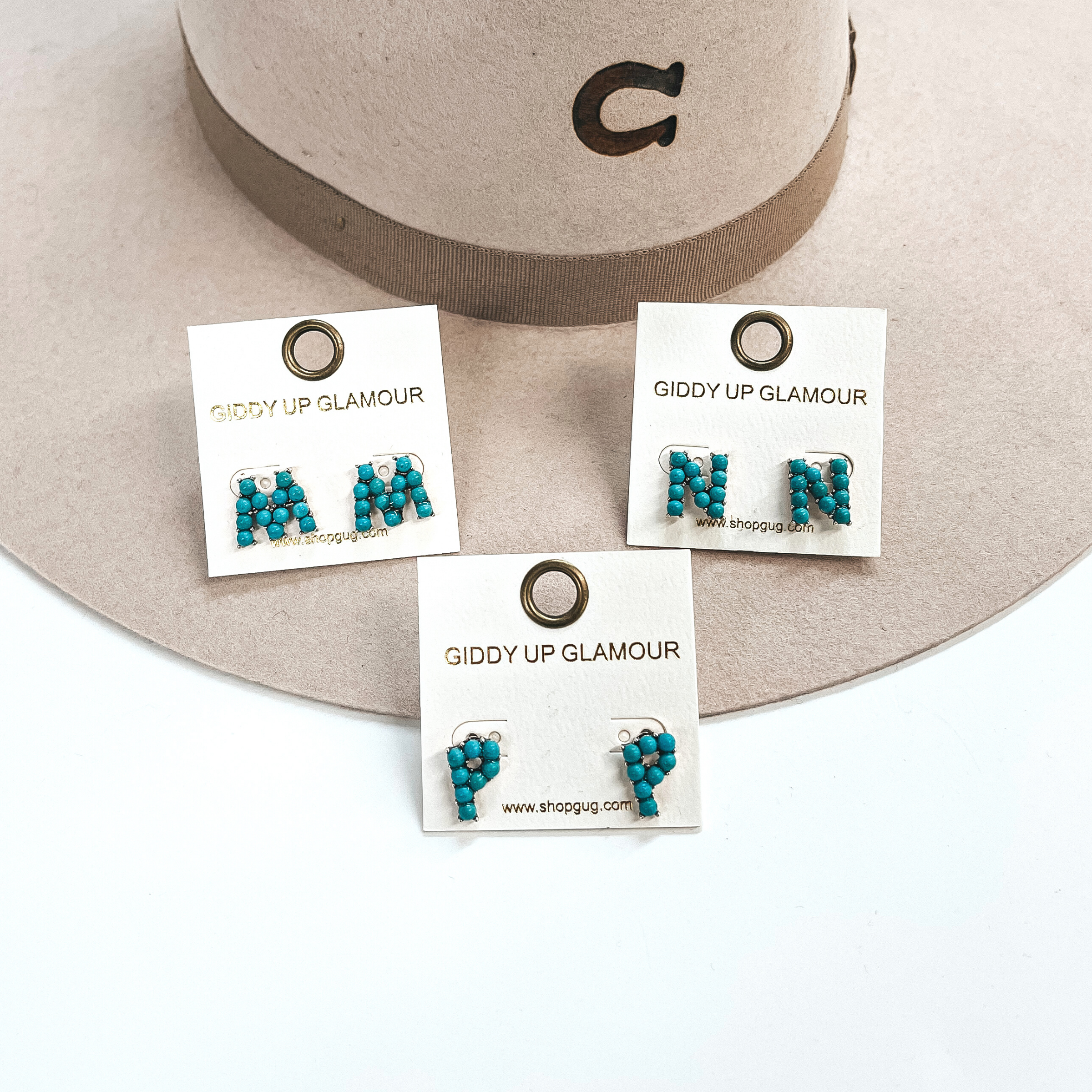 These are three pair of initial earrings with small turquoise stones in  a silver setting. From left to right; M,P,N. These earrings are placed  on a white Giddy Up Glamour card, they are laying on a beige, felt,  Charlie 1 Horse hat and on a white background.