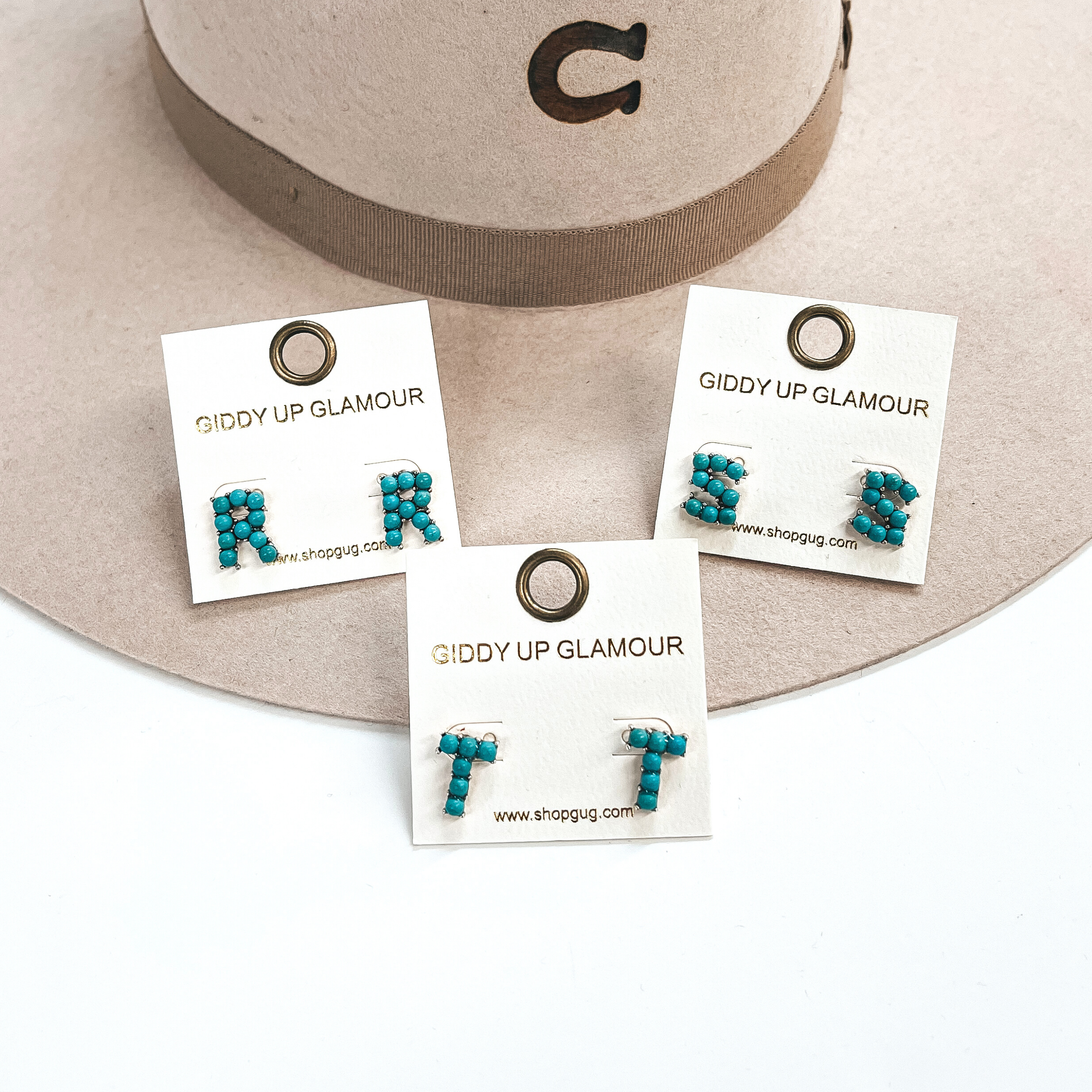 These are three pair of initial earrings with small turquoise stones in  a silver setting. From left to right; R,T,S. These earrings are placed  on a white Giddy Up Glamour card, they are laying on a beige, felt,  Charlie 1 Horse hat and on a white background.