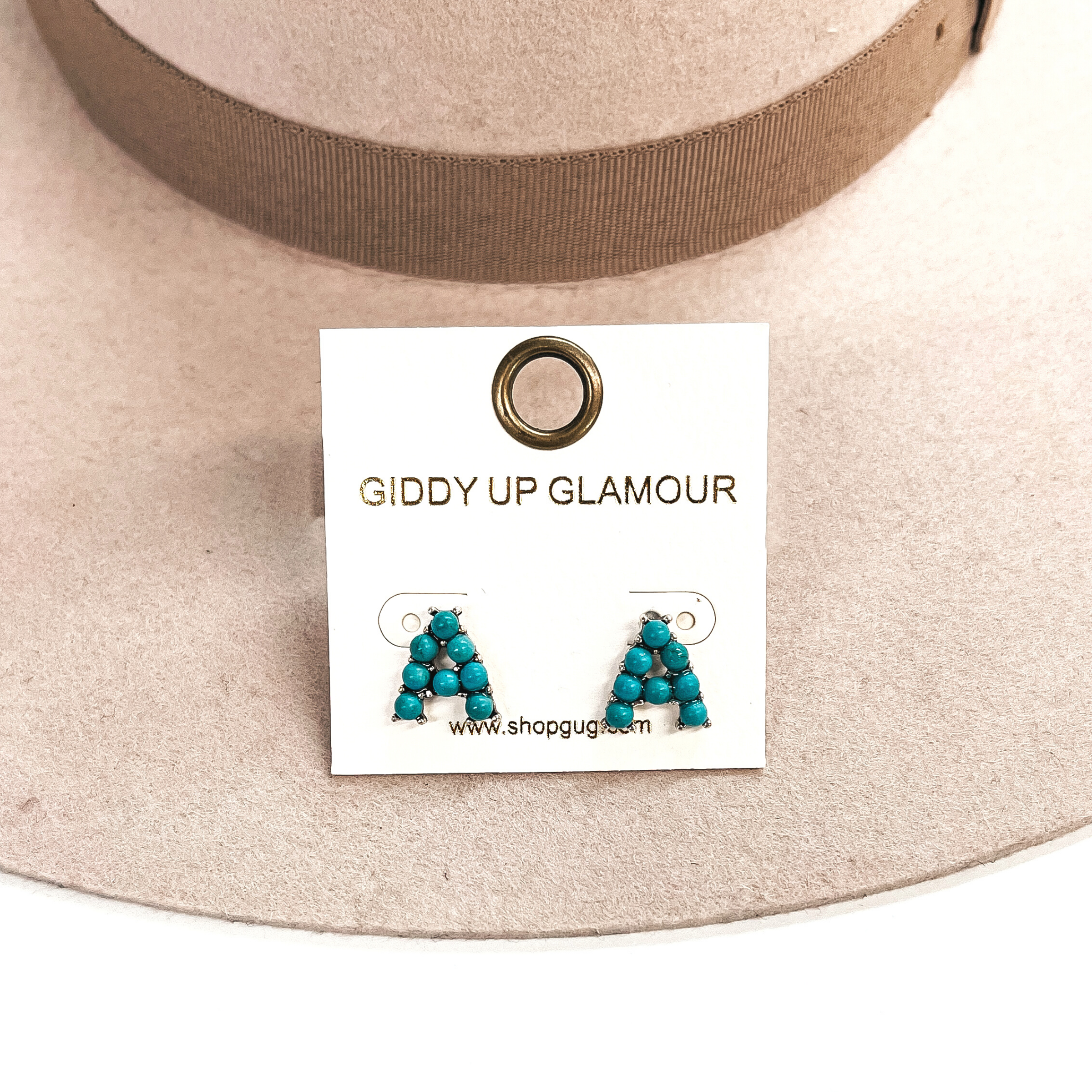 This is a pair of earrings with the initial, A, with small turquoise  stones and in a silver setting. These earrings are placed in a white,  Giddy Up Glamour card, this is taken on a beige felt hat and on a white  background.