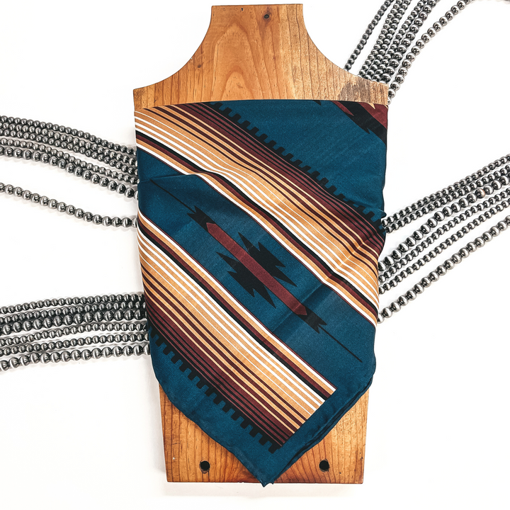 This is a  dark teal silk wild rag in aztec print with dark maroon, tan, and  white colors. This wild rag is taken on a brown necklace board and on a  white background with silver Navajo pearls in the back as decor.