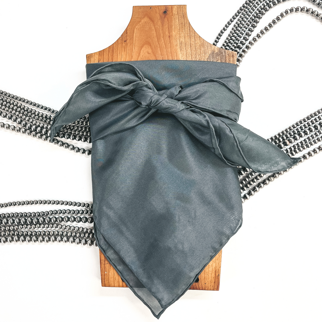 This is a charcoal grey  silk wild rag, this wild rag is placed on a brown necklace board. This wild rag is taken on a white background and with silver Navajo pearls in the back as decor.