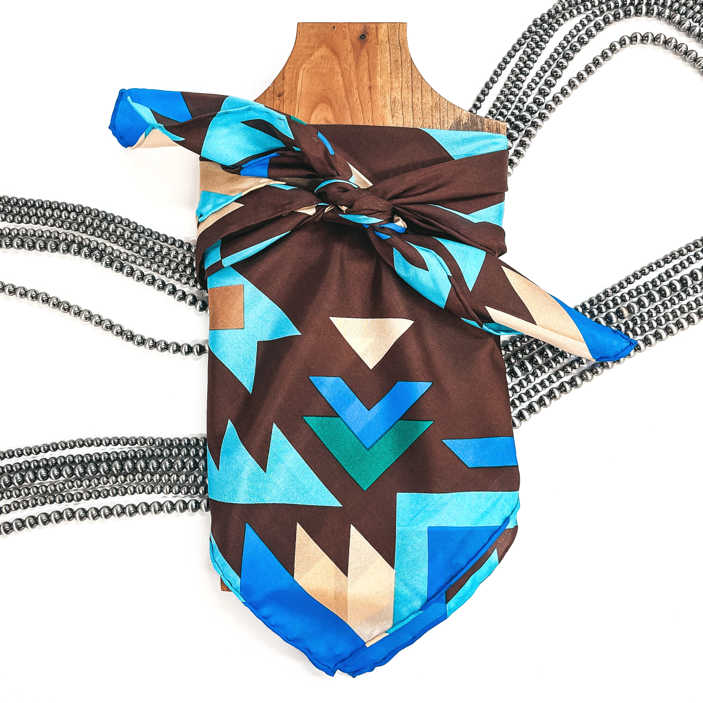 This is a silk wild rag in tribal print in brown, beige, turquoise, and blue. This wild rag is taken on a brown necklace board and on a white  background with silver Navajo pearls in the back as decor.