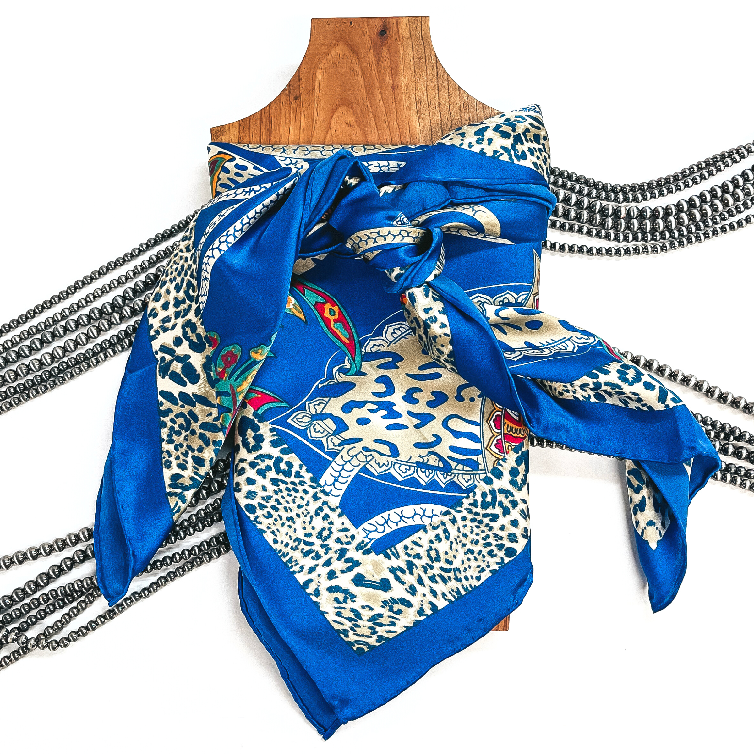 This is a silk wild rag in royal blue with charmuese print in various other prints such as leopard and paisley. The colors are beige, champagne gold, yellow, teal, green, pink, and orange. This wild rag is taken on a brown necklace board and white background with silver Navajo pearls in the back as decor.