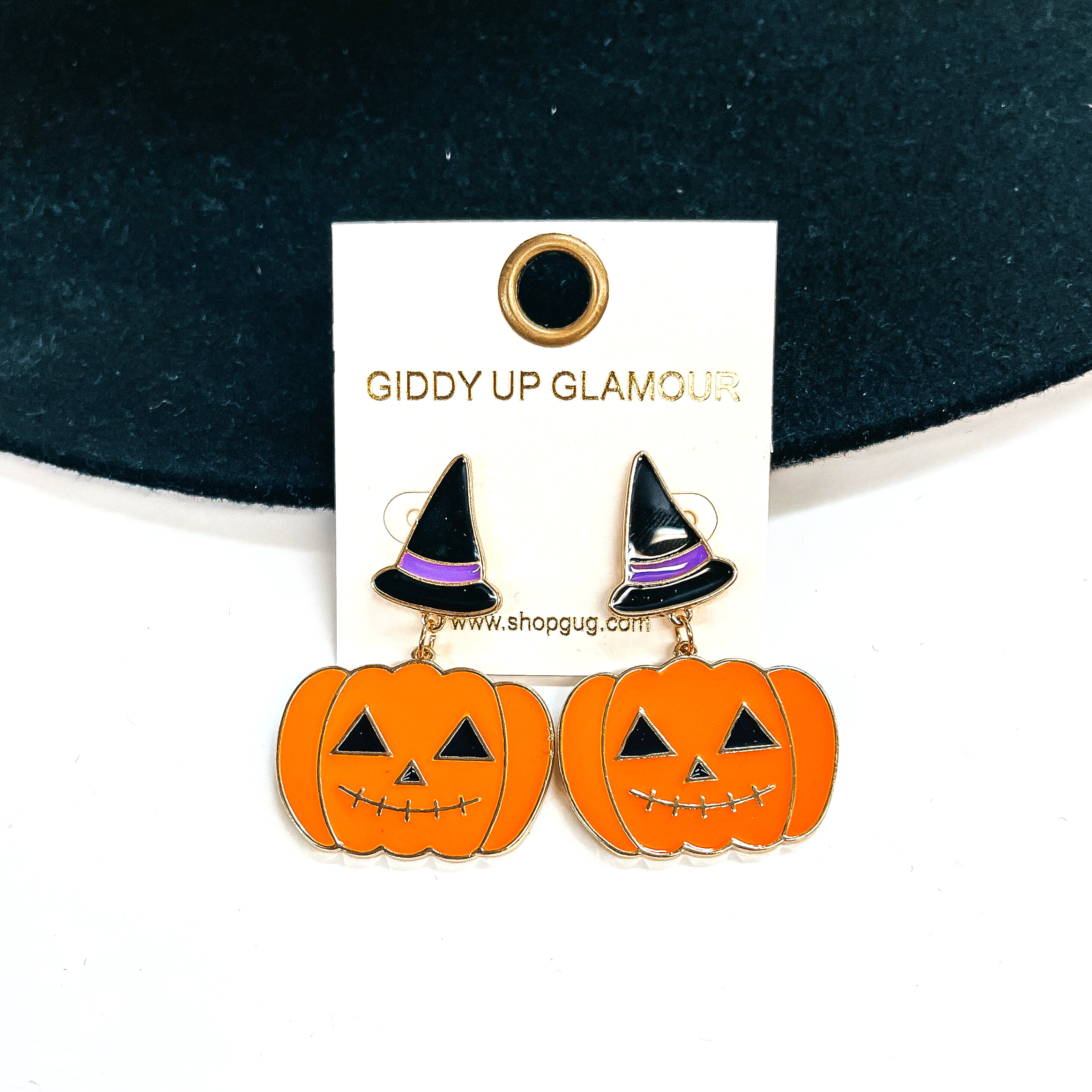 Black witch hat post earrings with a purple band, the drop has an orange  Jack O'Lantern in a gold setting. These earrings are taken leaning on a  black felt hat and on a white background.