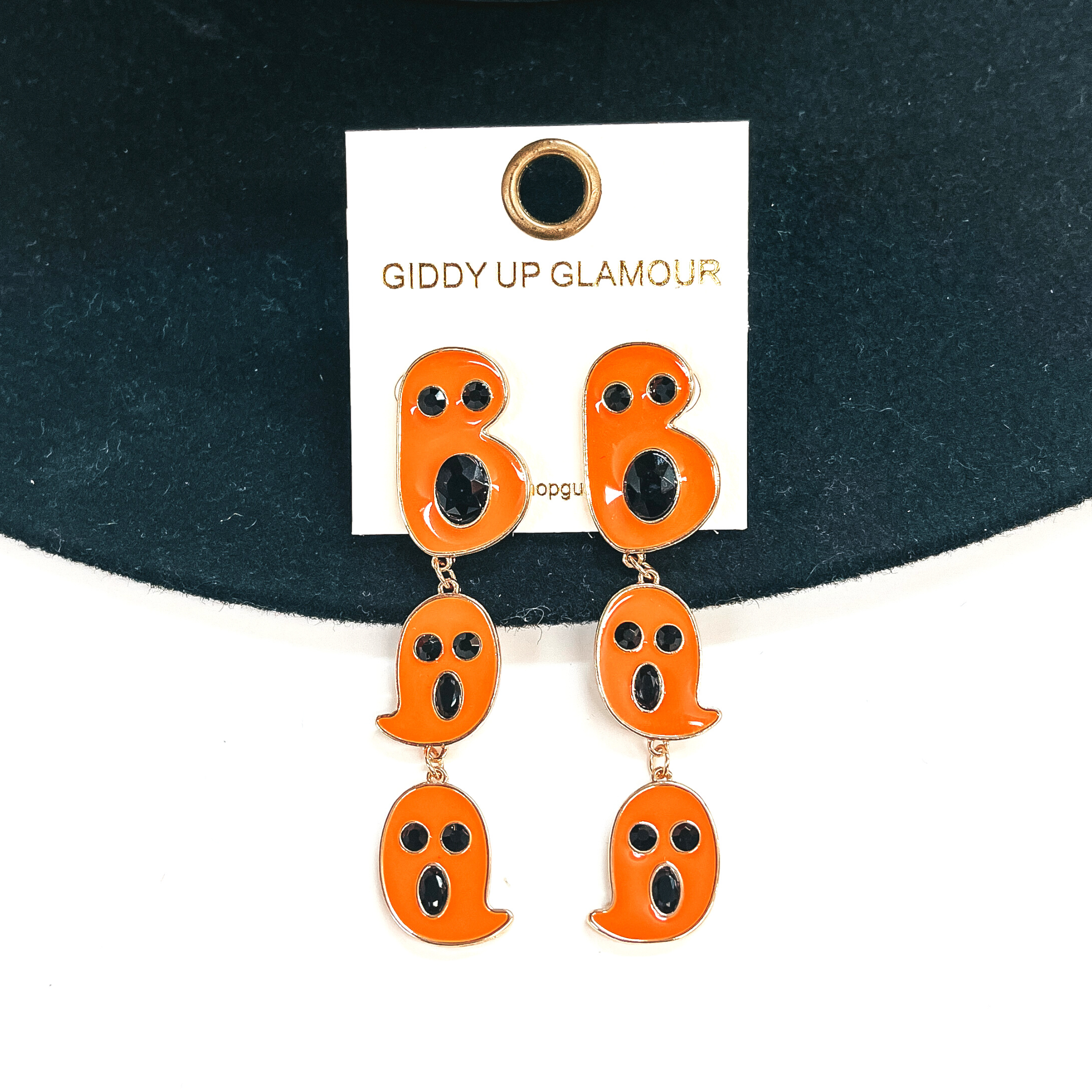 These are ghost shaped tiered drop earrings that spell out, Boo. They are in a gold setting and have black crystals as eyes and mouth, these are orange.  These earrings are taken on a black felt hat brim and on a white background.