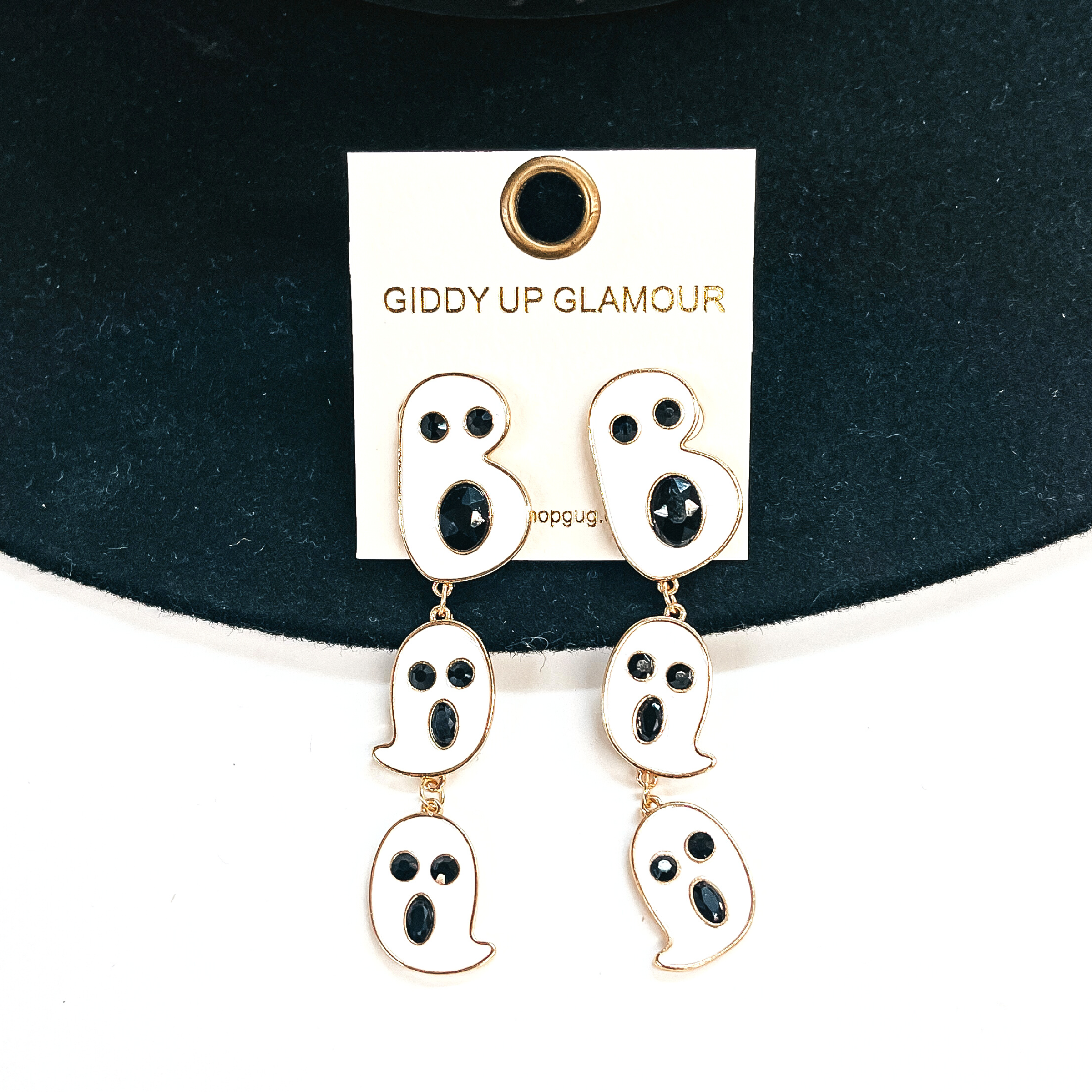 These are ghost shaped tiered drop earrings that spell out, Boo. They are in a gold setting and have black crystals as eyes and mouth, these are white.  These earrings are taken on a black felt hat brim and on a white background.