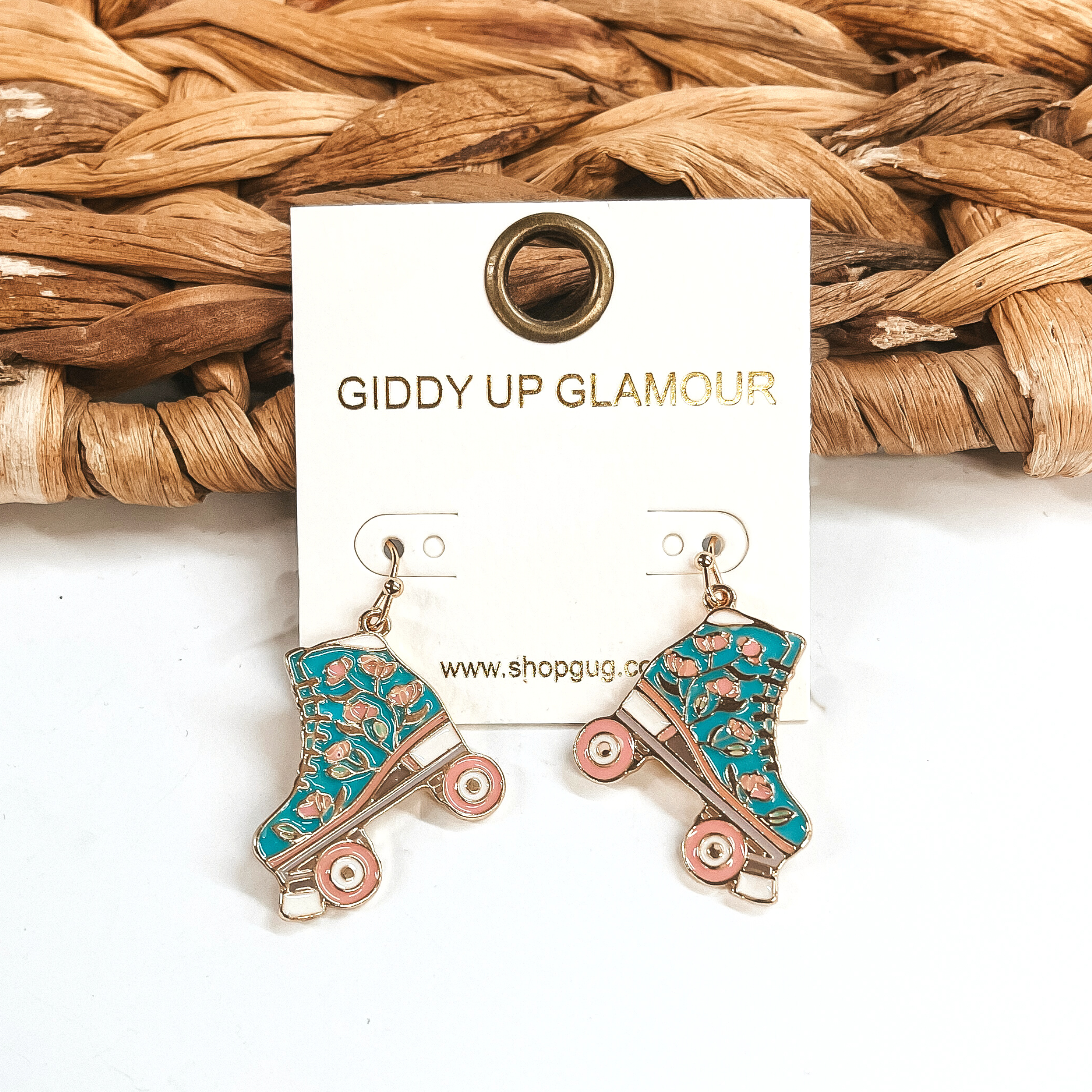 These are turquoise rollerskate earrings in a gold setting. There is a mix of different  colors such as turquoise, light pink and white. There are light pink flowers on the  rollerskate.  These earrings are taken leaning up against a brown woven slate and on a white  background.