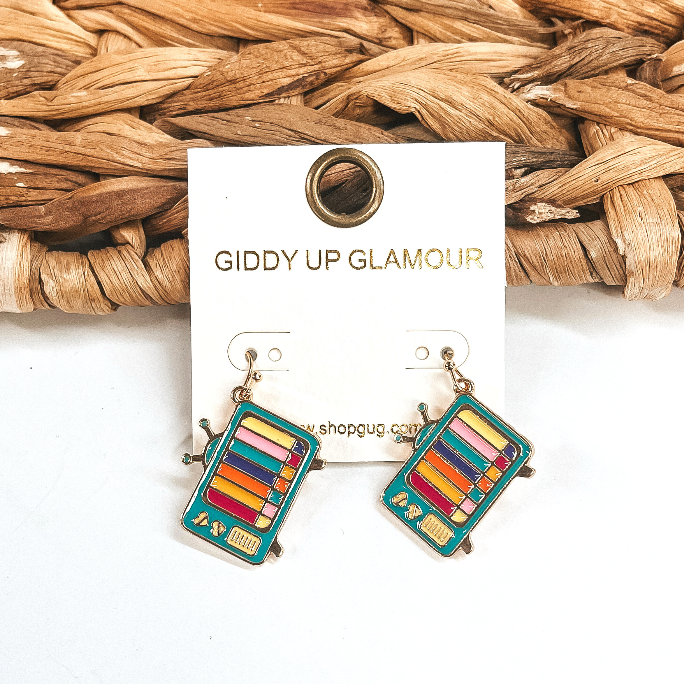 These are television shape earrings in teal blue-green and multicolor lines on the screen in a  gold setting. There are small hearts in the side of the TV.  These earrings are taken leaning up against a brown woven slate and on a white  background.