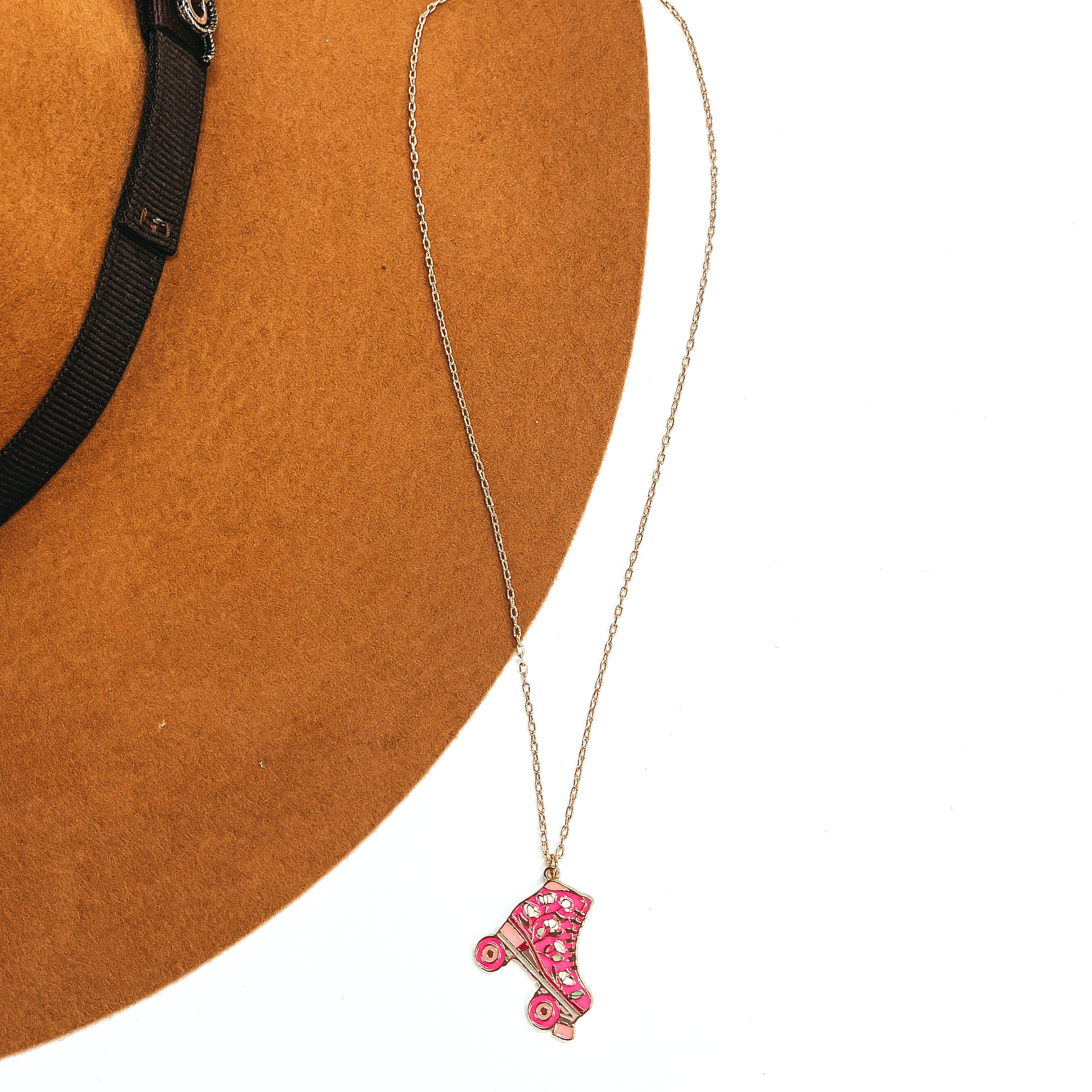 Gold thin chain necklace with a pink rollerskate pendant. The pink rollerskate has  white floral design in the center with different shades of pink. This necklace is taken  laying on a camel-brown, felt hat brim and on a white background.