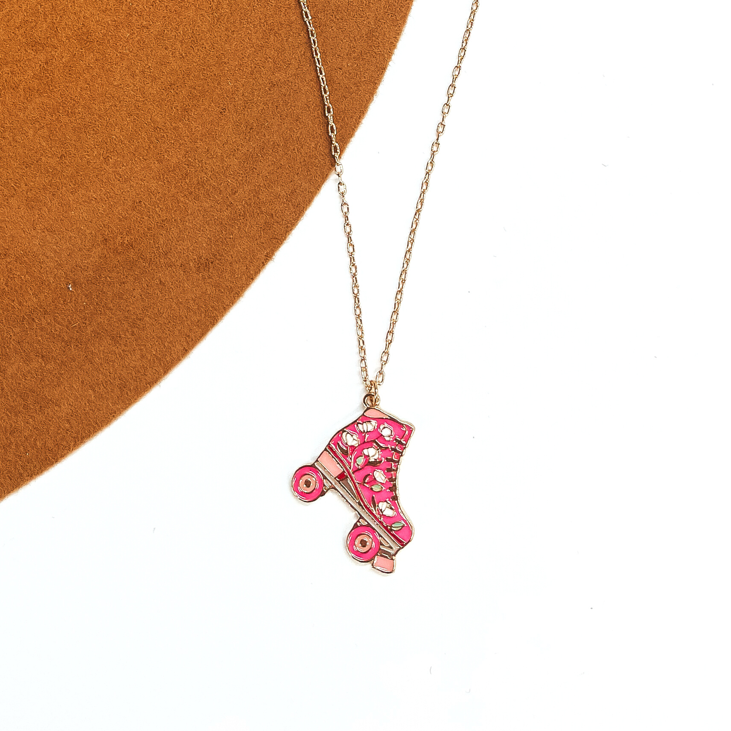 Gold thin chain necklace with a pink rollerskate pendant. The pink rollerskate has  white floral design in the center with different shades of pink. This necklace is taken  laying on a camel-brown, felt hat brim and on a white background.