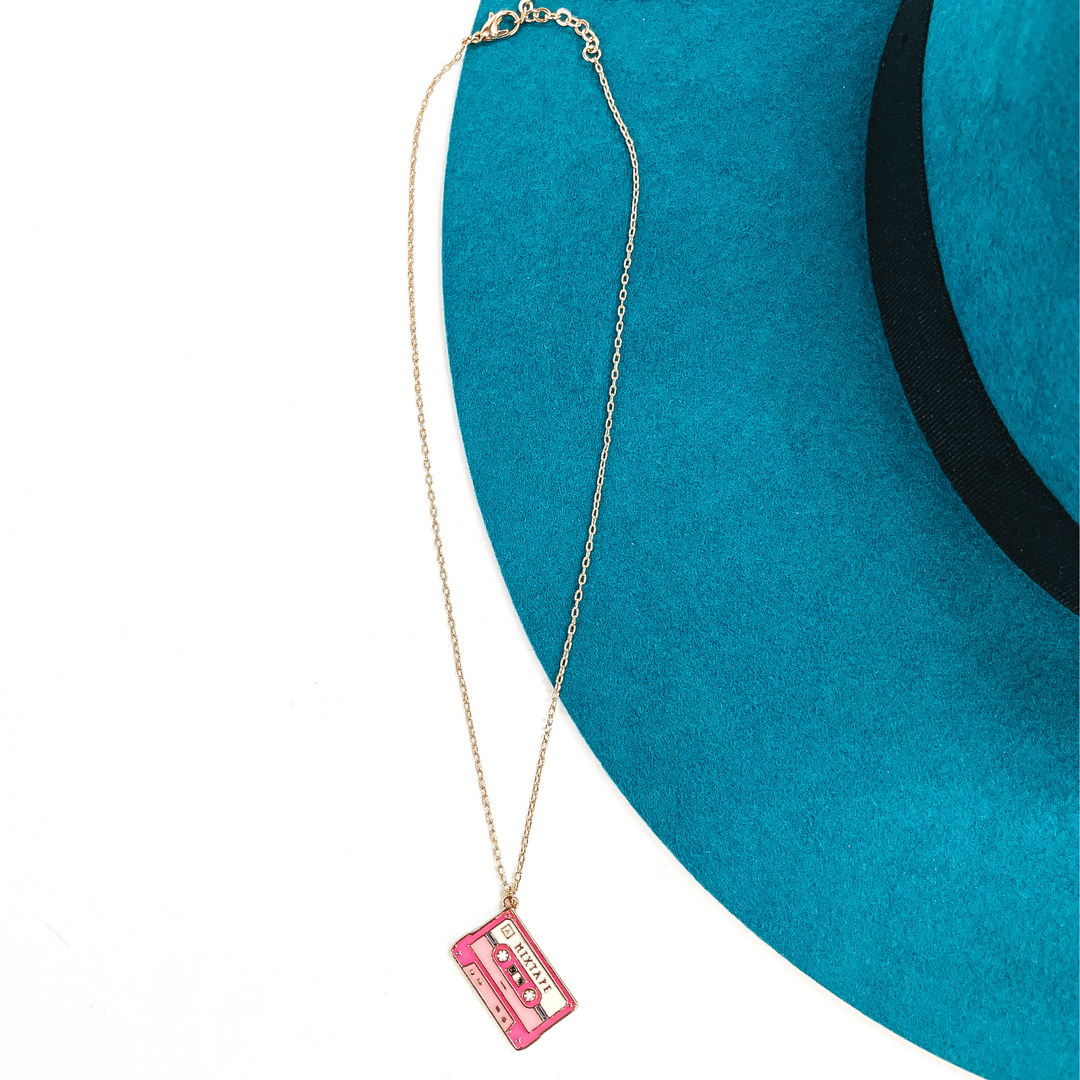 Gold thin chain necklace with a pink cassette tape pendant. The cassette pendant  has different shades of pink and with gold tone embellishments. This necklace  is laying on a white background and a teal felt hat brim.