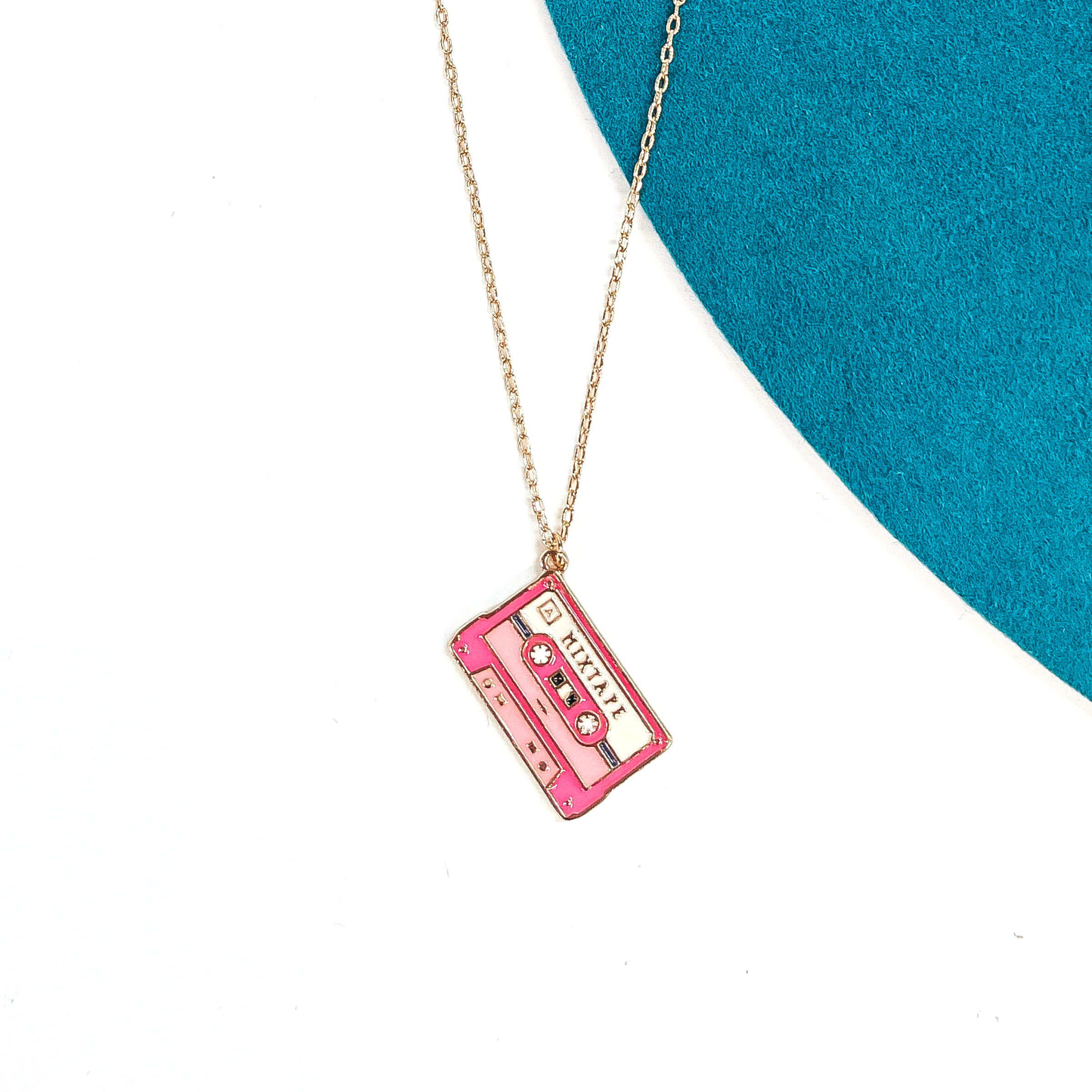 Gold thin chain necklace with a pink cassette tape pendant. The cassette pendant  has different shades of pink and with gold tone embellishments. This necklace  is laying on a white background and a teal felt hat brim.