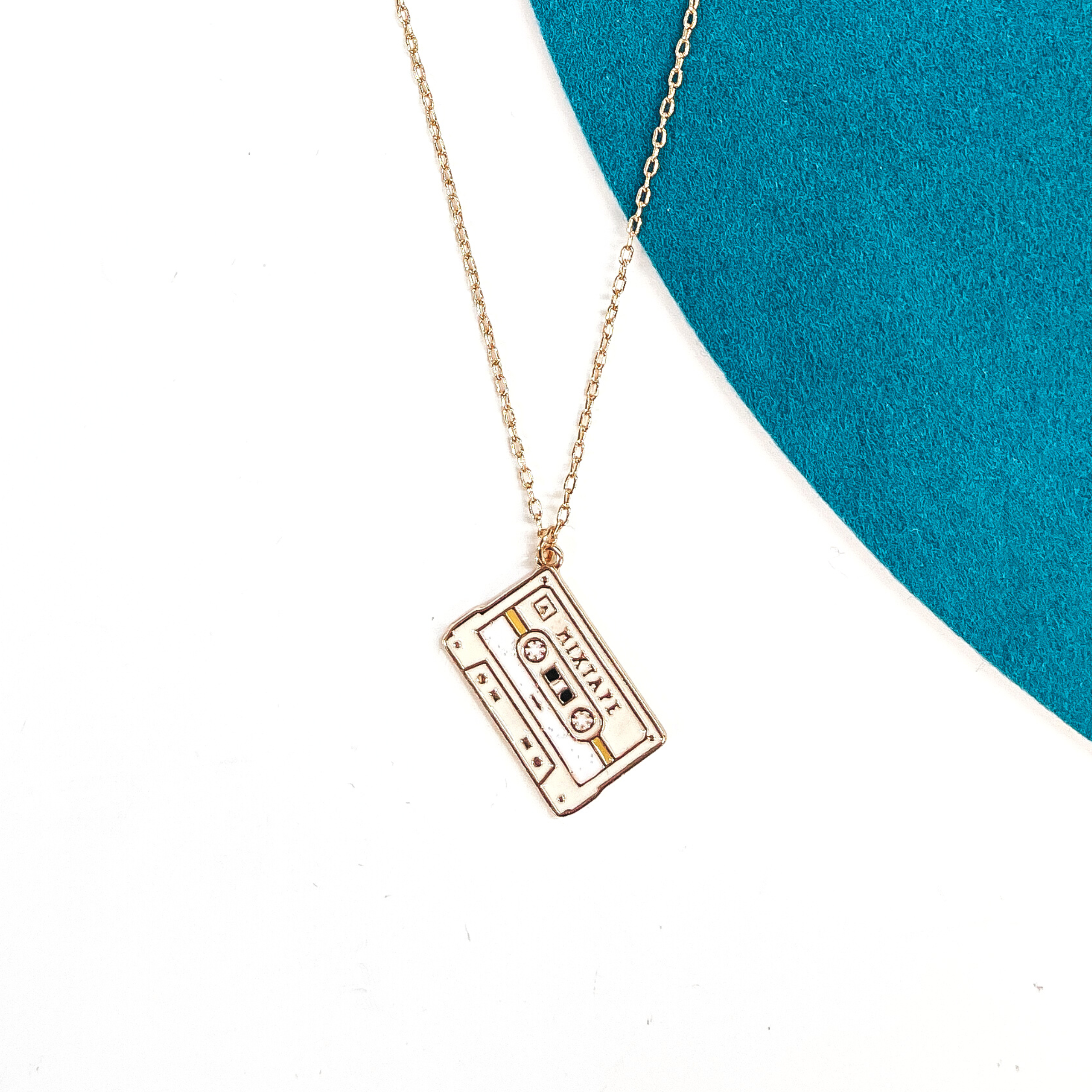 Gold thin chain necklace with an ivory cassette tape pendant. The cassette pendant  has a bit of glitter in the center and other tones of white and tan. This necklace  is laying on a white background and a teal felt hat brim.