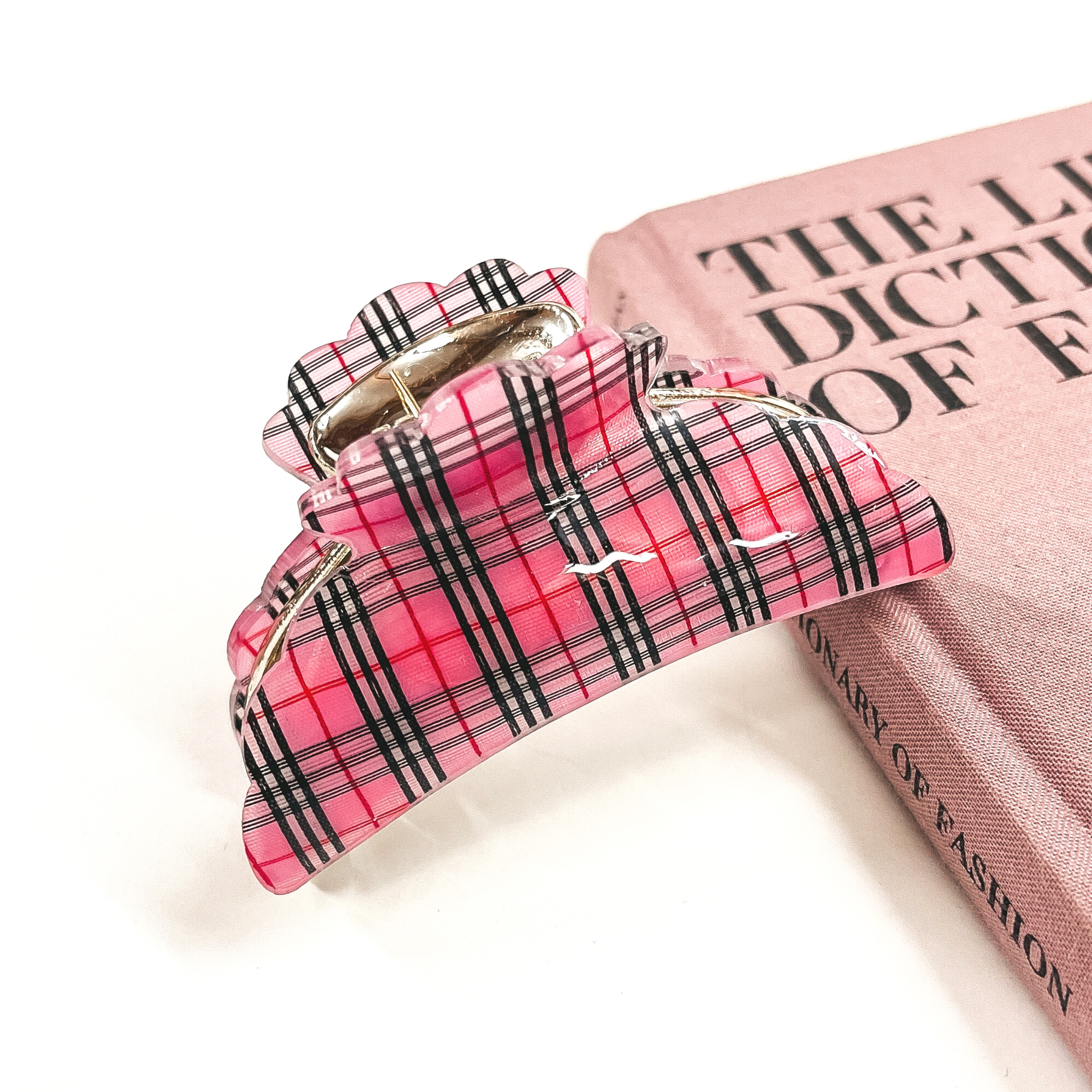 This is a gold and pink plaid print hair clip laying on the side of a pink book and white background.