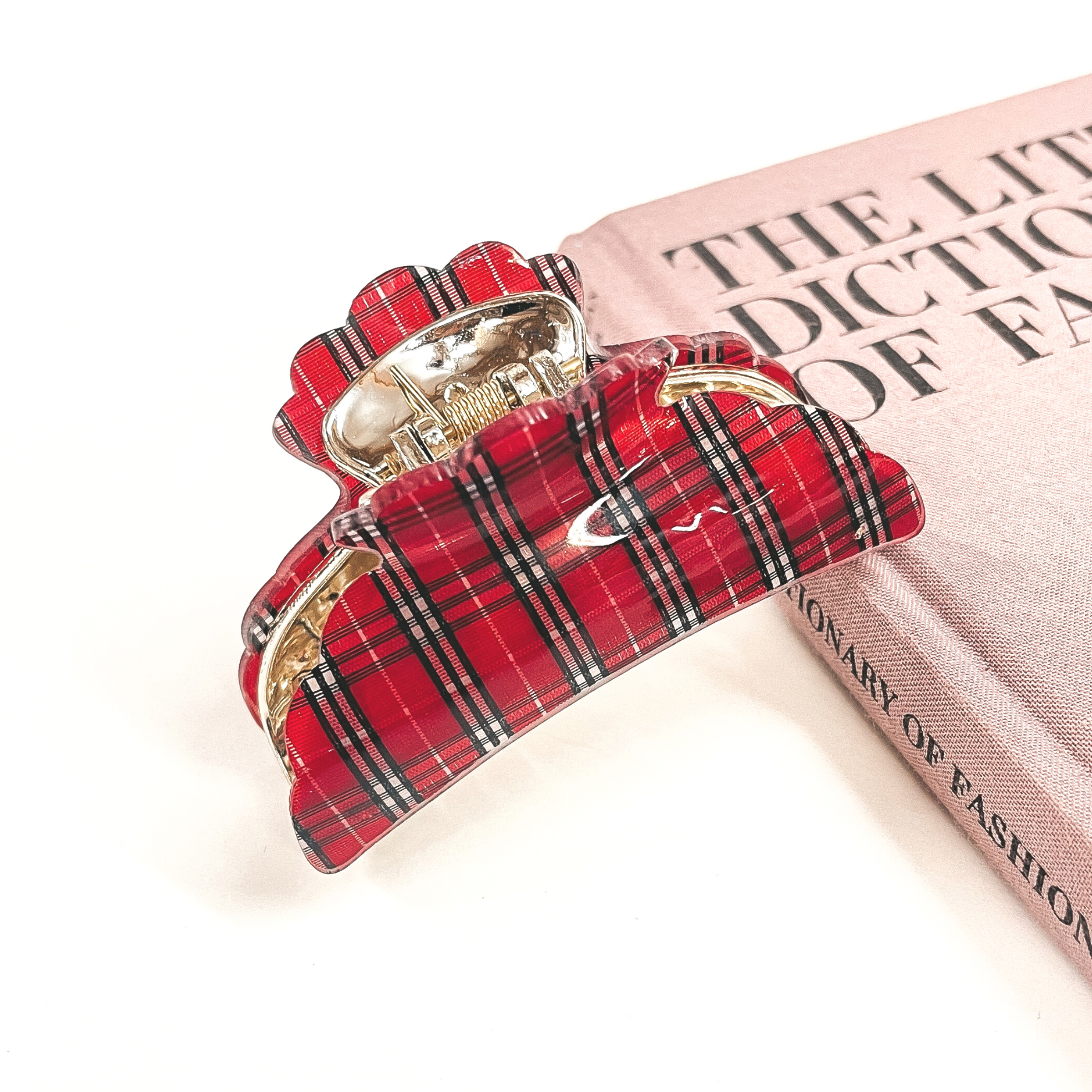 This is a gold and red plaid print hair clip laying on the side of a pink book and white background.