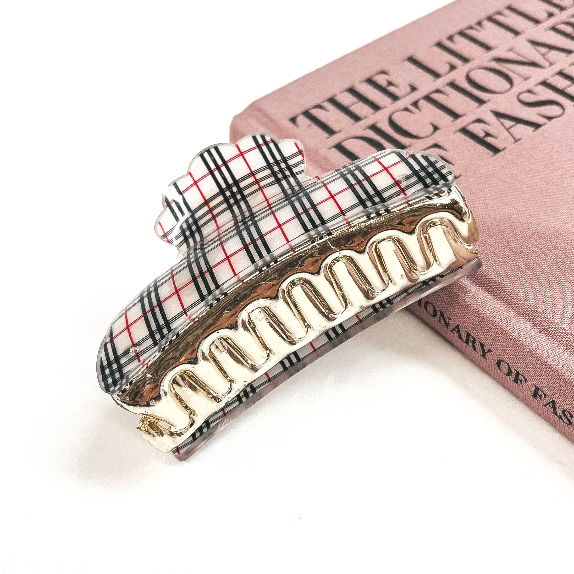 This is a gold and white plaid print hair clip laying on the side of a pink book and white background.
