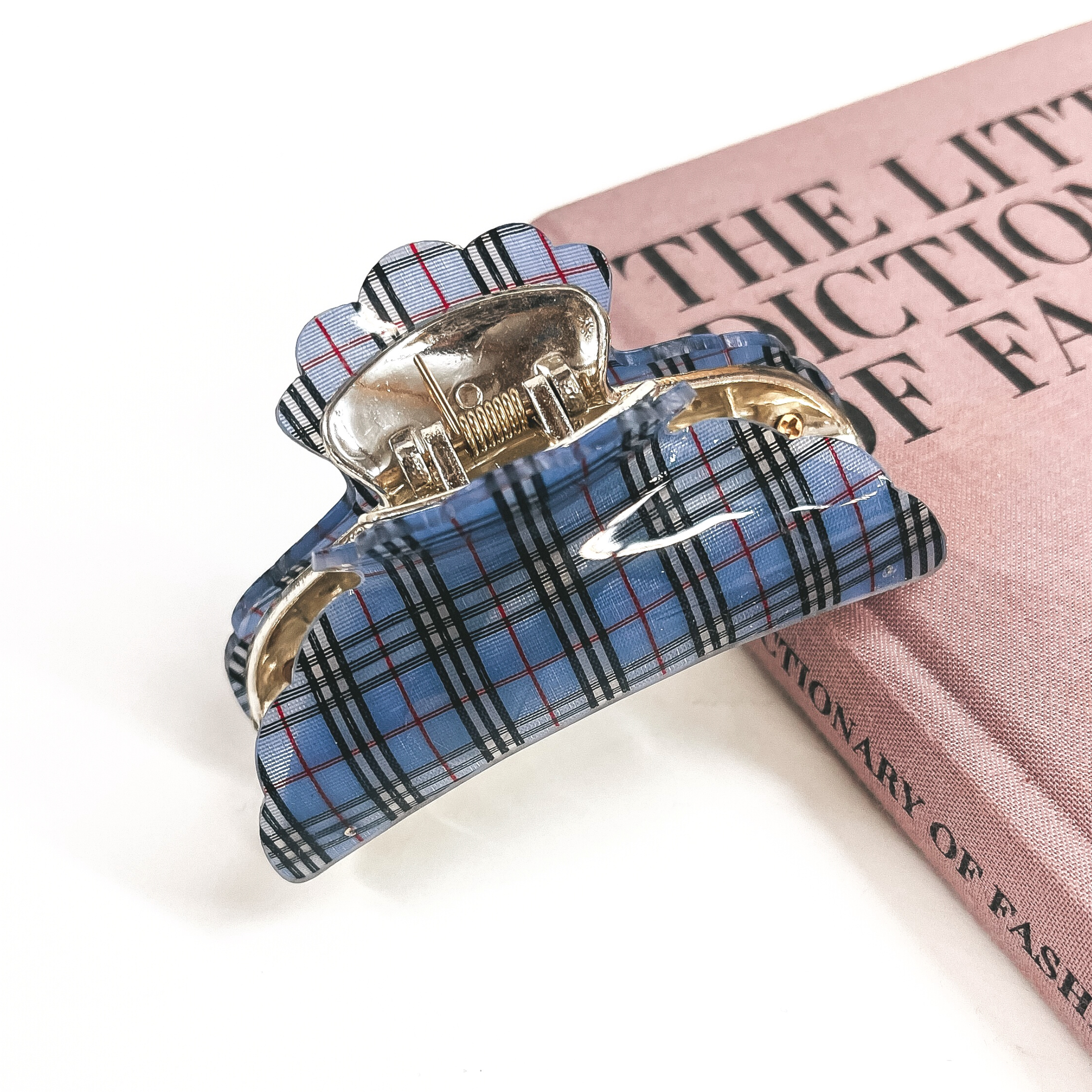 This is a gold and blue plaid print hair clip laying on the side of a pink book and white background.