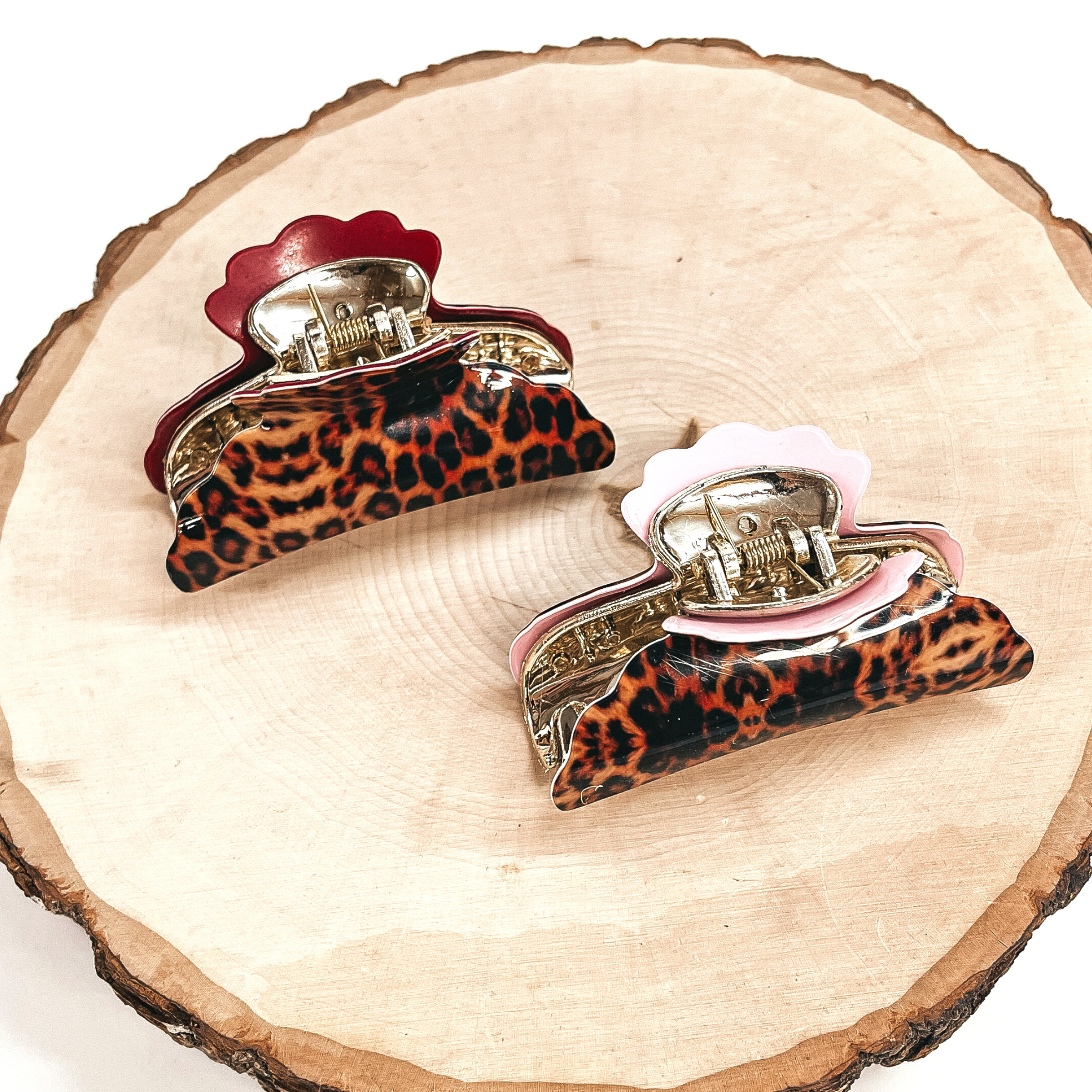 There are two orange leopard print hair clips, one is in red and the other is light  pink with gold tone inlay. Both of these hair clips are taken on a slab of wood and  a white background.