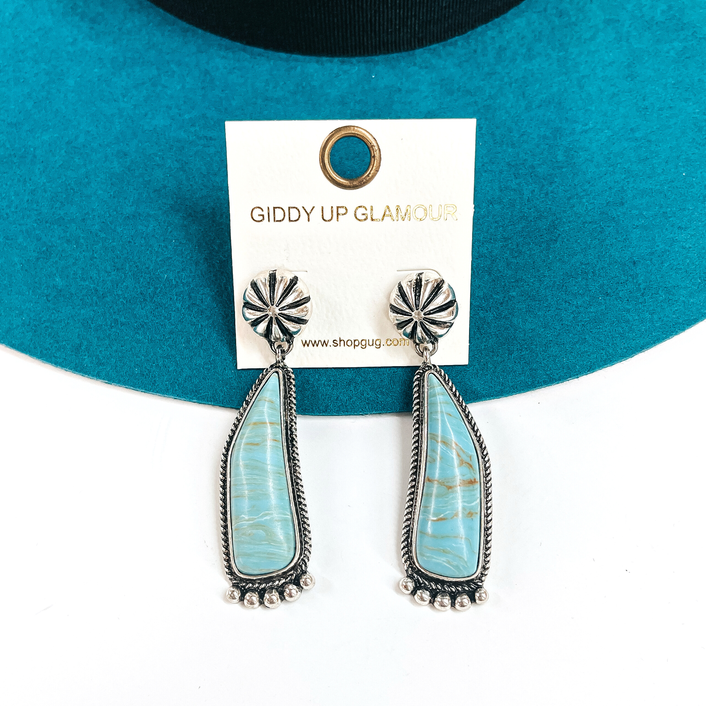 These are silver conch post earrings with a turquoise-marble stone drop earrings in a  silver setting. They have silver details in the bottom and they ae slightly curved.  These earrings are taken laying on a teal felt hat and on a white background.
