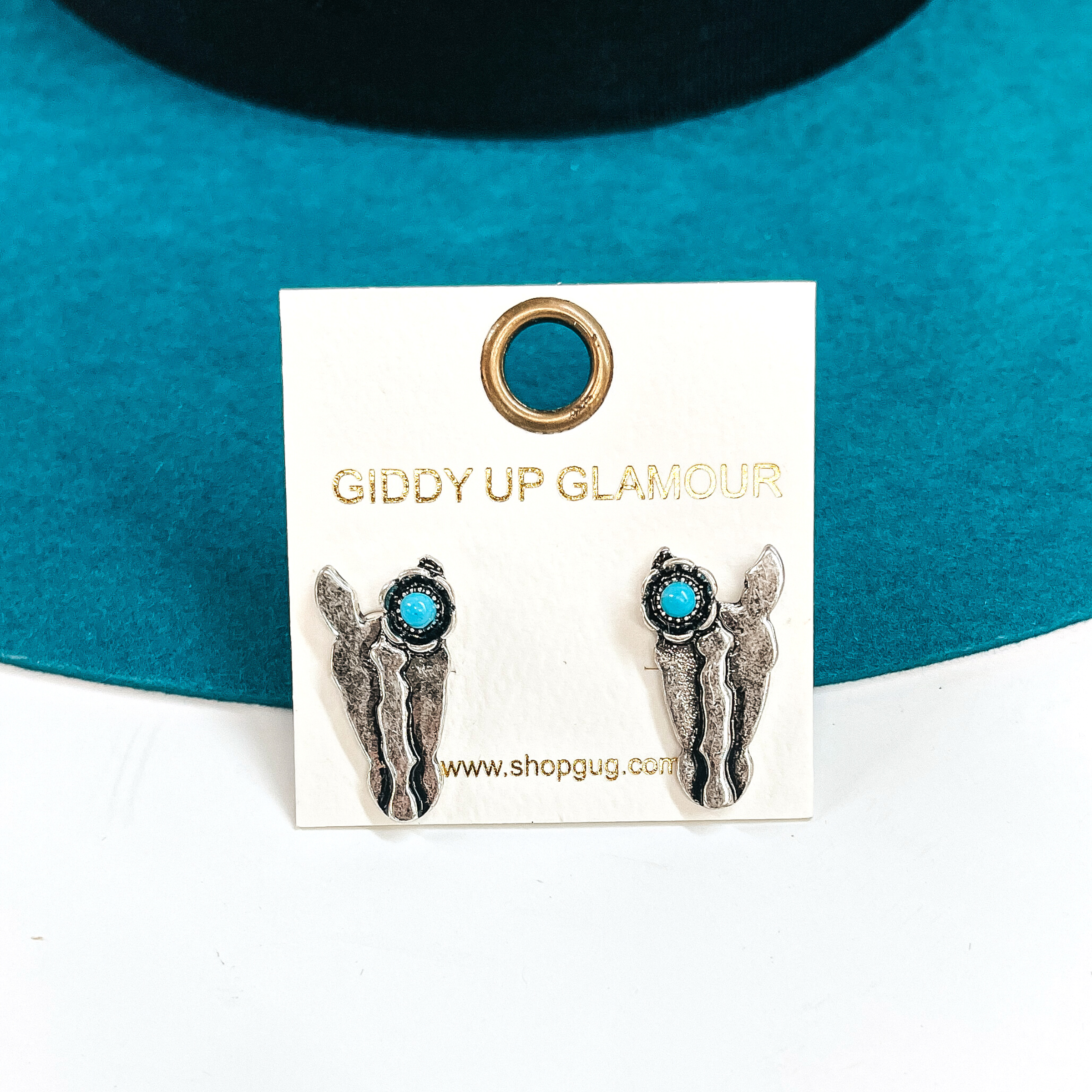 These are silver stud earrings in a horse head shape with a small silver detailed flower  on the ear. The flower has a small fake turquoise stone.  These earrings are taken laying on a teal felt hat and on a white background.