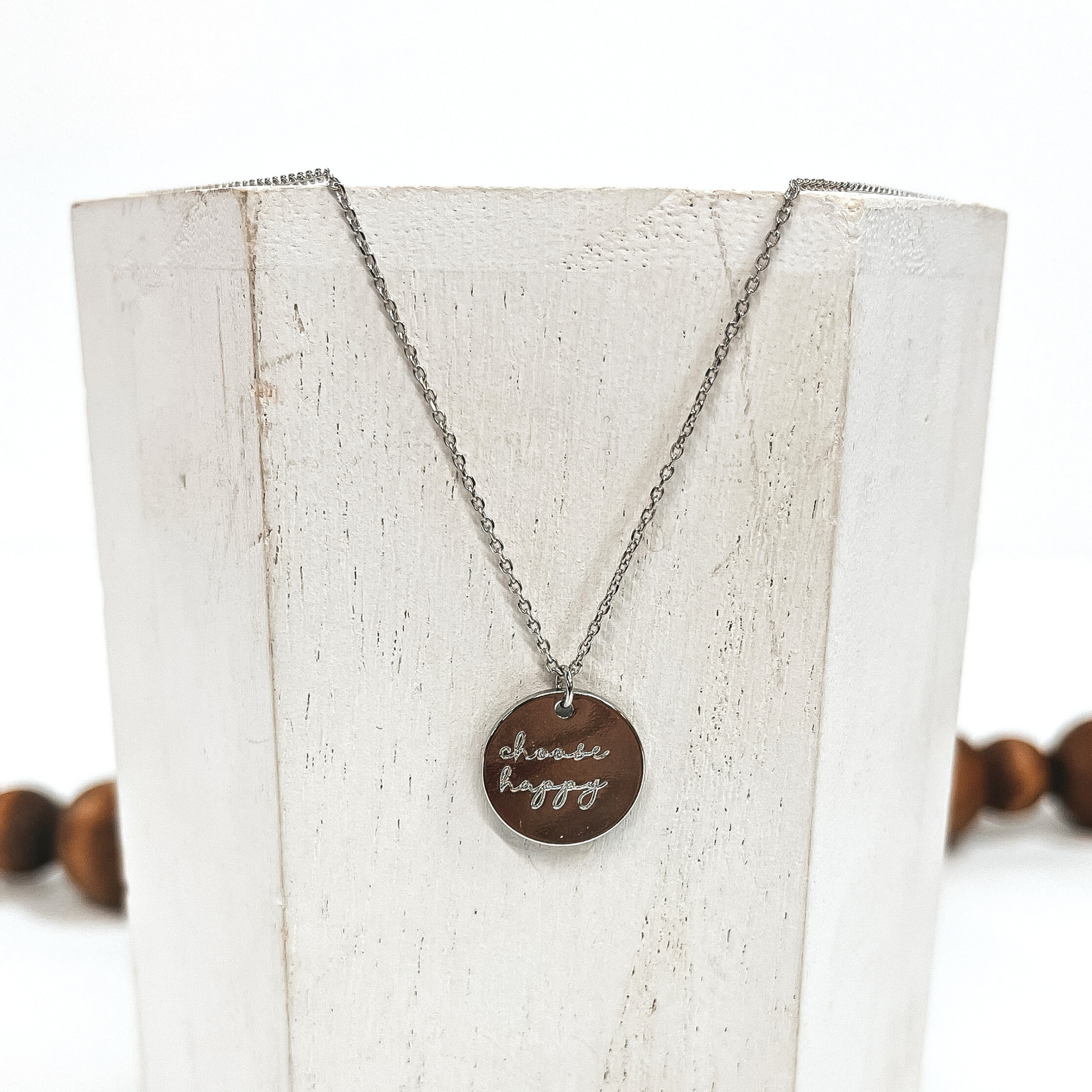 This is a thin silver necklace with a silver pendant that says, 'choose happy'.  This necklace is taken on a white block and on a white background with brown  beads in the back as decor.