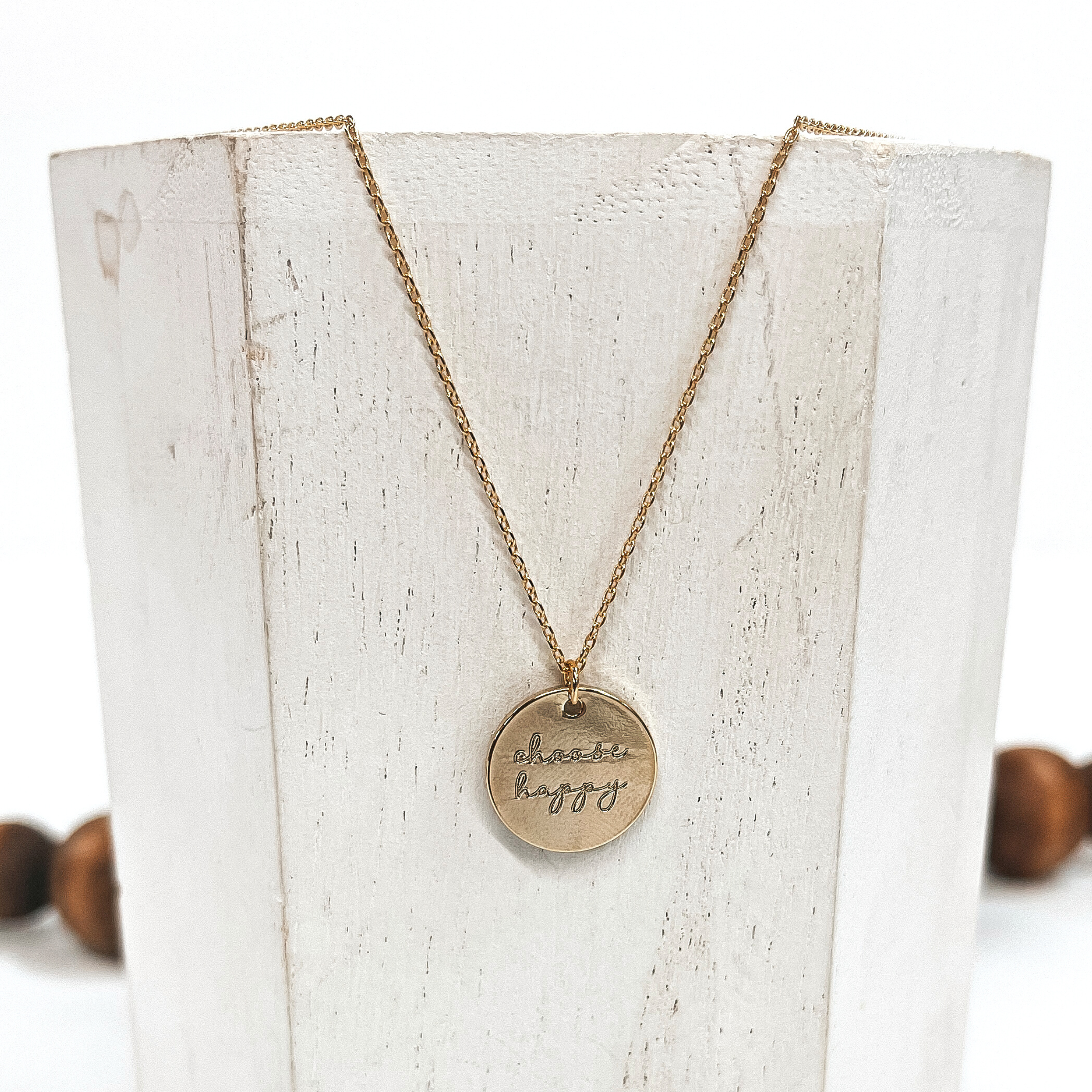 This is a thin gold necklace with a gold pendant that says, 'choose happy'.  This necklace is taken on a white block and on a white background with brown  beads in the back as decor.