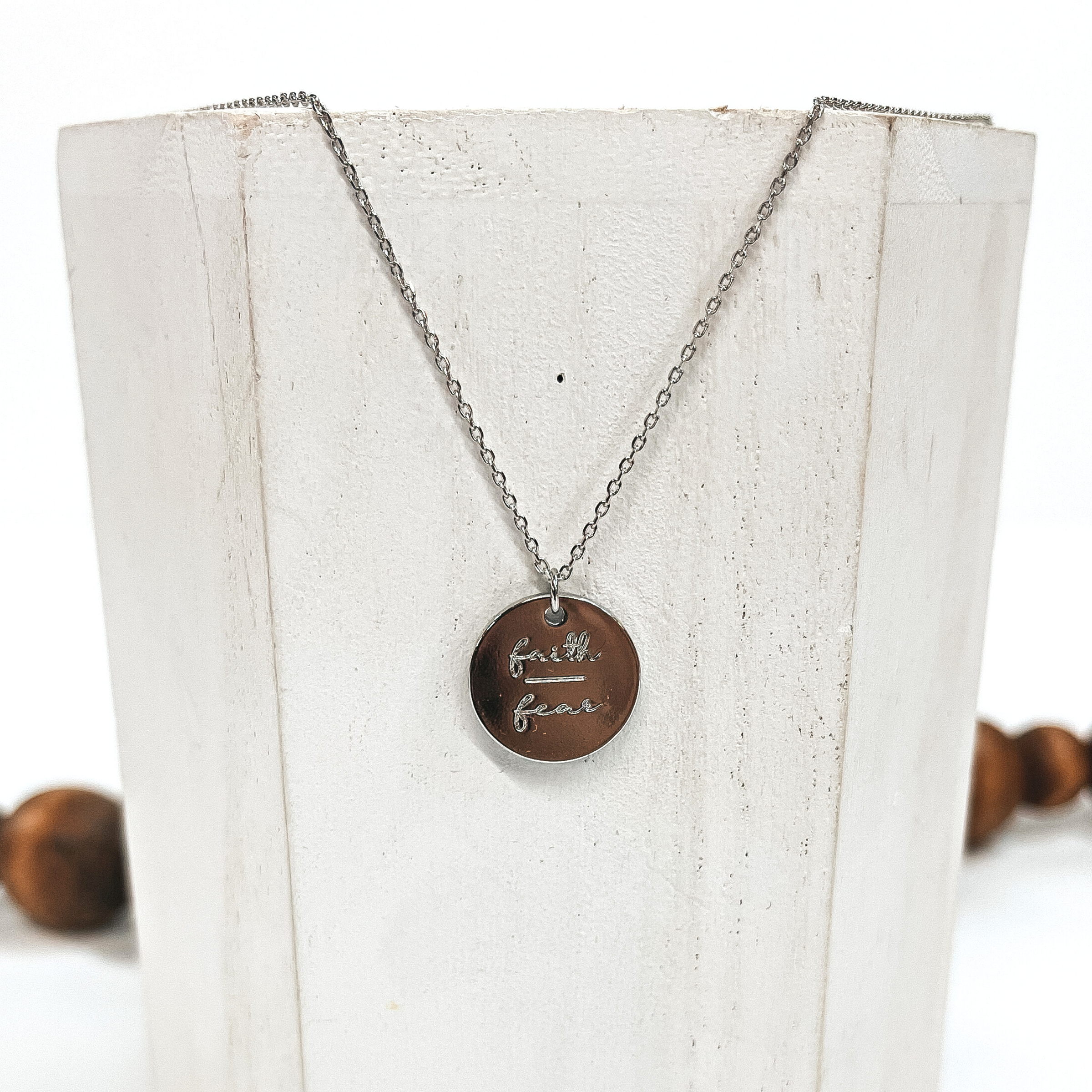 This is a thin silver necklace with a silver pendant that says, 'Faith over fear'.  This necklace is taken on a white block and on a white background with brown  beads in the back as decor.
