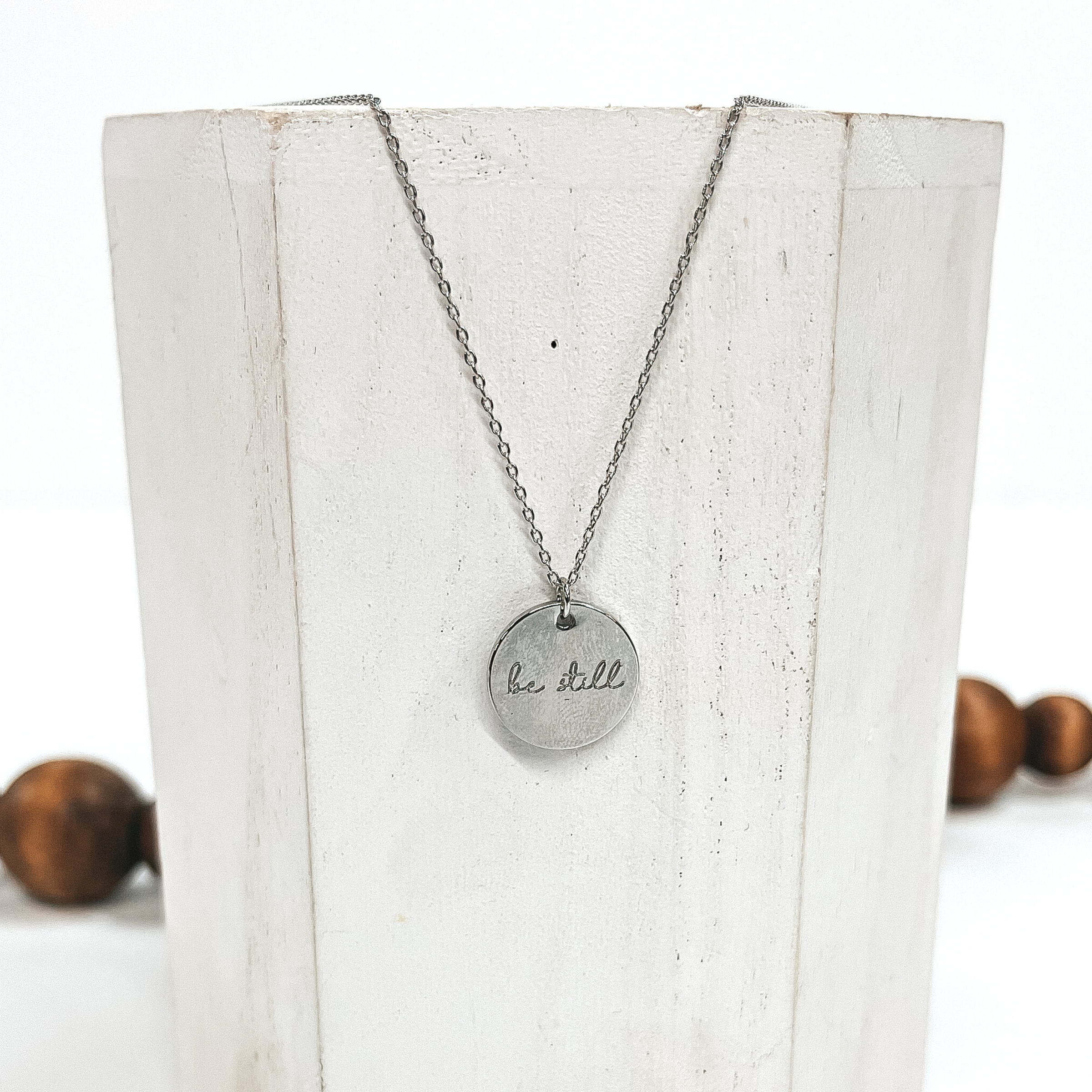 This is a thin silver necklace with a silver pendant that says, 'be still'.  This necklace is taken on a white block and on a white background with brown  beads in the back as decor.