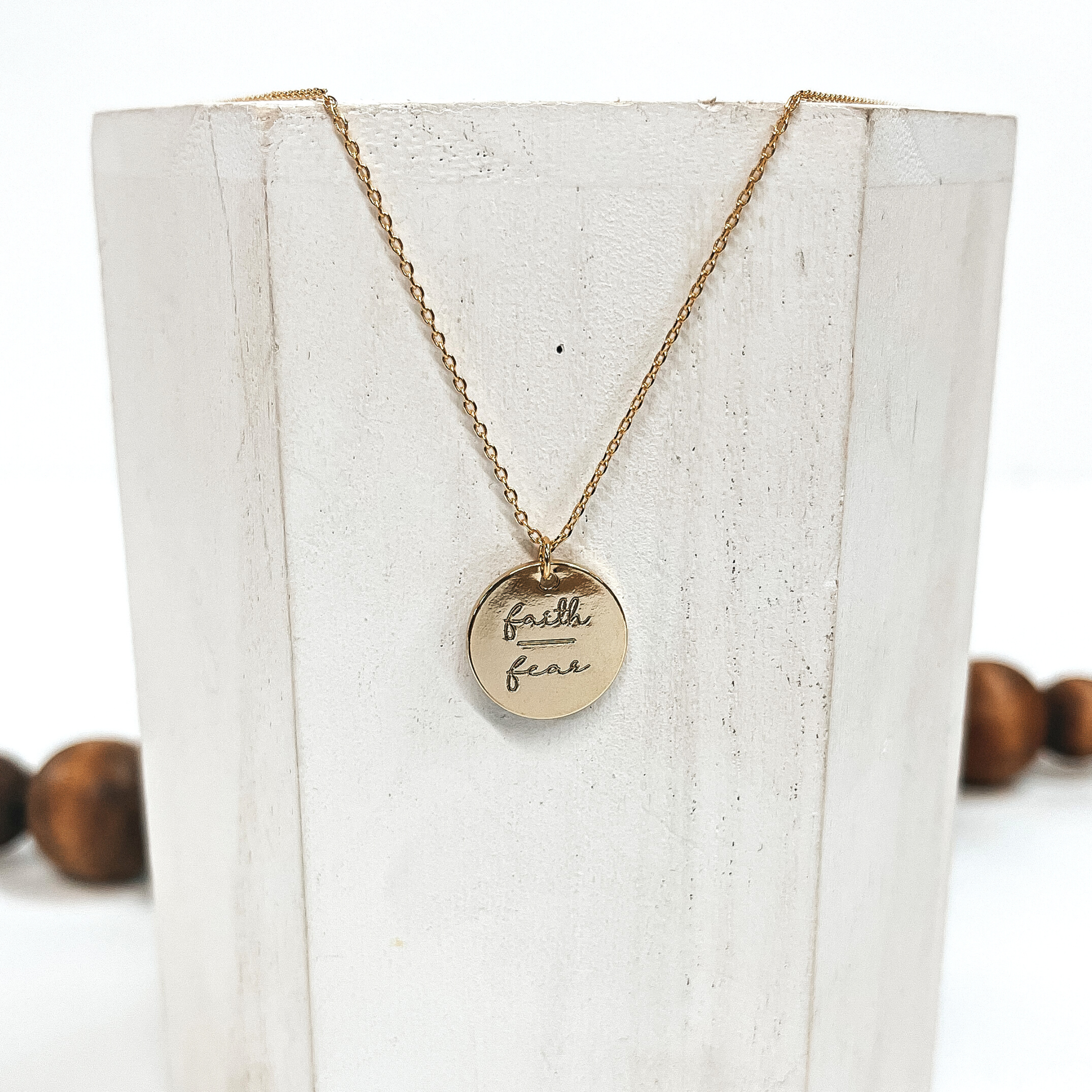 This is a thin gold necklace with a gold pendant that says, 'Faith over fear'.  This necklace is taken on a white block and on a white background with brown  beads in the back as decor.