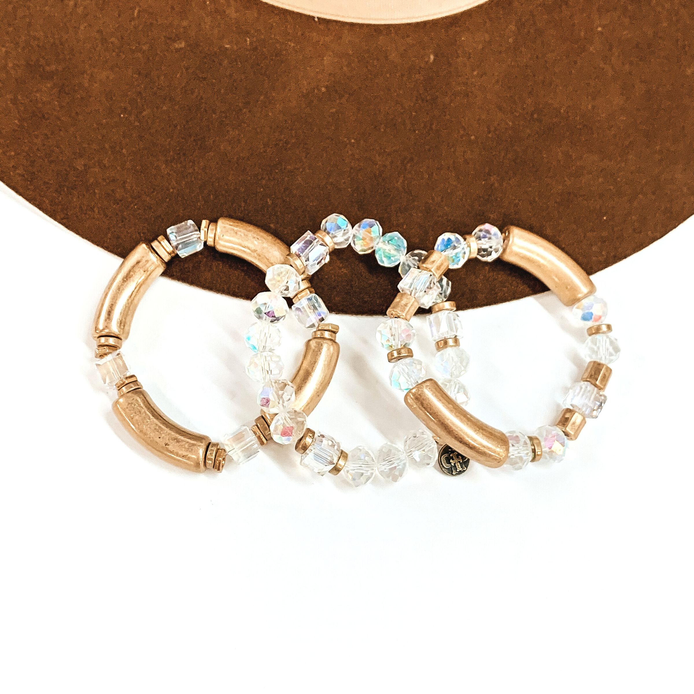This is a set of three bangle bracelets in gold and ab clear crystals in different  shapes. From left to right; gold tubes with square ab clear crystals, the middle  bracelet has a mix of circle and square ab clear crystals. The right bracelet  has two gold tubes, circle ab clear crystals, and gold square beads. These  bracelets are taken laying on a dark brown felt hat and on a white background.