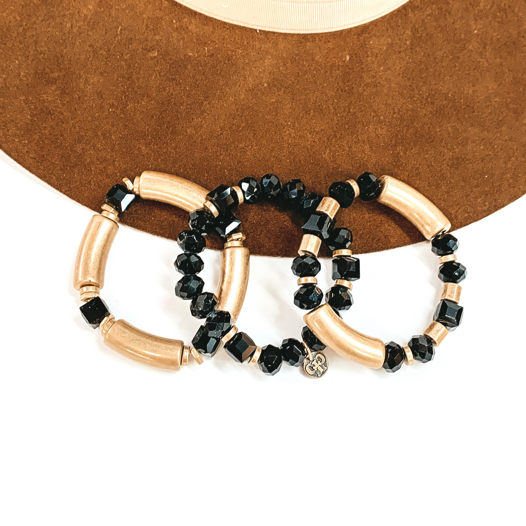 This is a set of three bangle bracelets in gold and black crystals in different  shapes. From left to right; gold tubes with square black crystals, the middle  bracelet has a mix of circle and square black crystals. The right bracelet  has two gold tubes, circle black crystals, and gold square beads. These  bracelets are taken laying on a dark brown felt hat and on a white background.