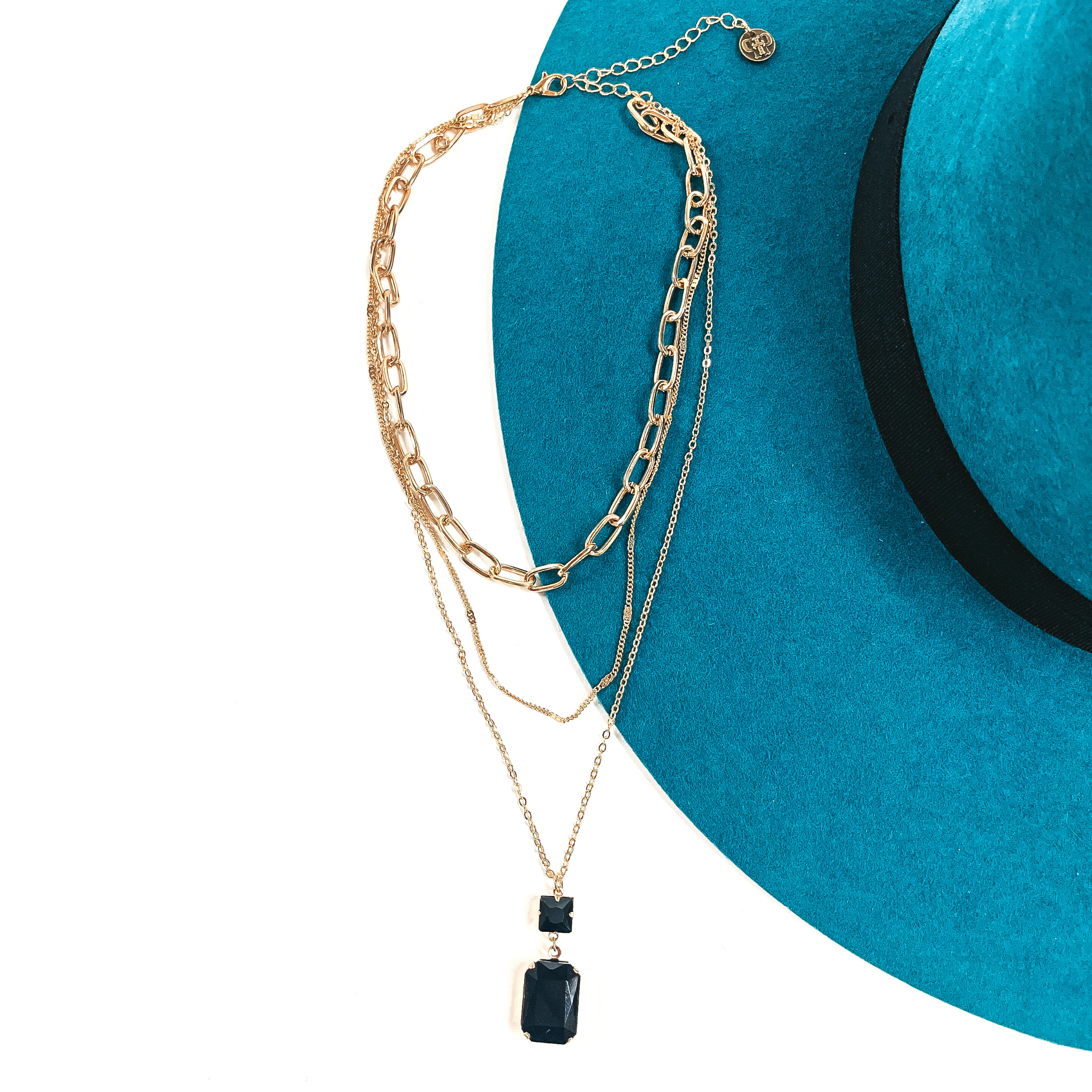 This is a three strand gold tone necklace with a black crystal pendant. There are  two thin strands and one thick chain link strand. The longest strand has a small  square black crystal connector and a rectangle black crystal. This necklace is taken  on a teal felt hat brim and on a white background.