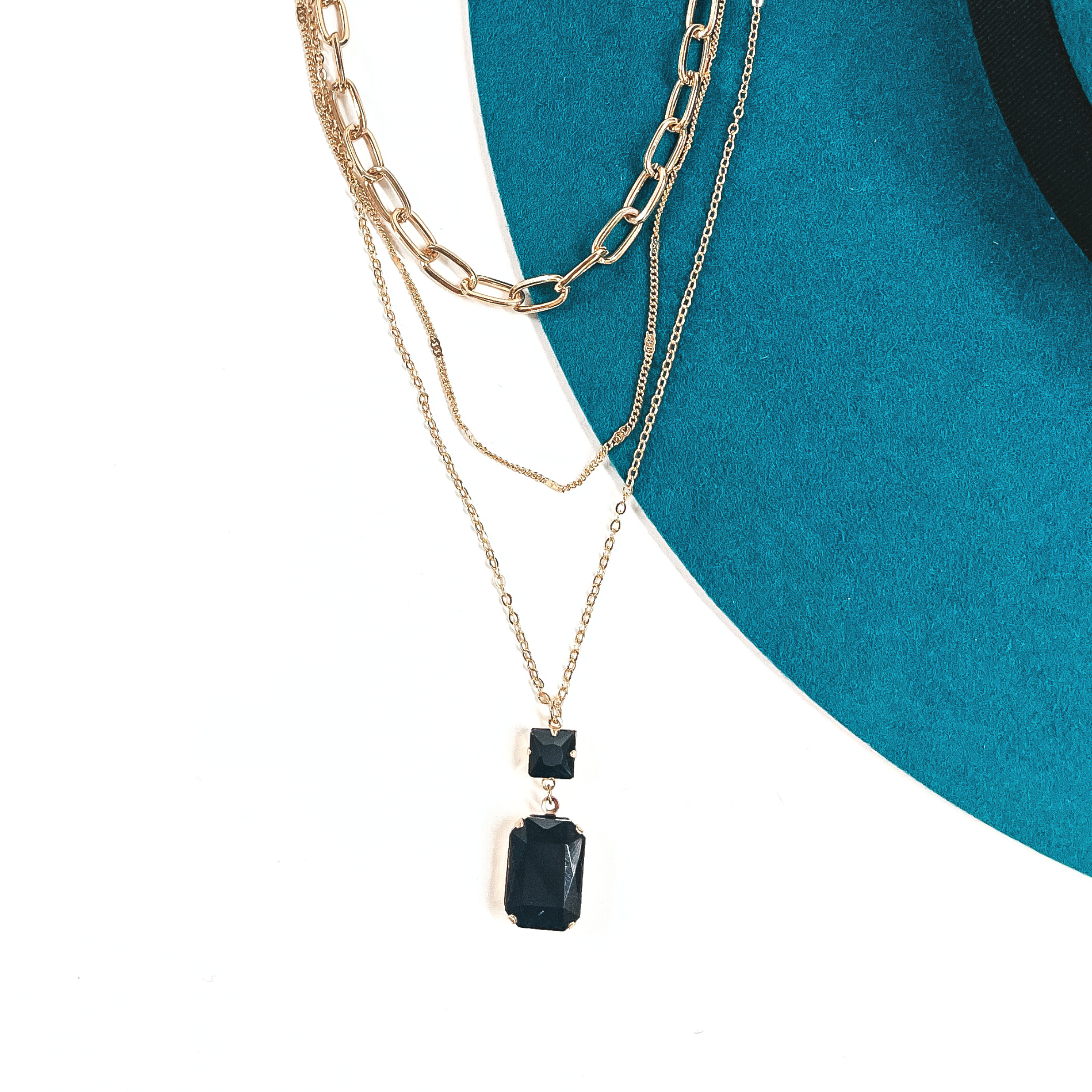 This is a three strand gold tone necklace with a black crystal pendant. There are  two thin strands and one thick chain link strand. The longest strand has a small  square black crystal connector and a rectangle black crystal. This necklace is taken  on a teal felt hat brim and on a white background.