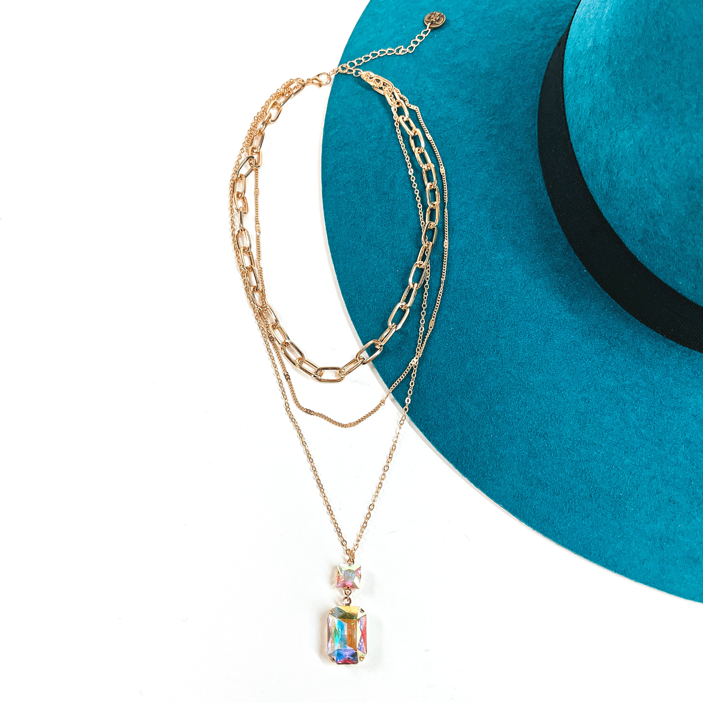 This is a three strand gold tone necklace with an AB crystal pendant. There are  two thin strands and one thick chain link strand. The longest strand has a small  square AB crystal connector and a rectangle AB crystal. This necklace is taken  on a teal felt hat brim and on a white background.