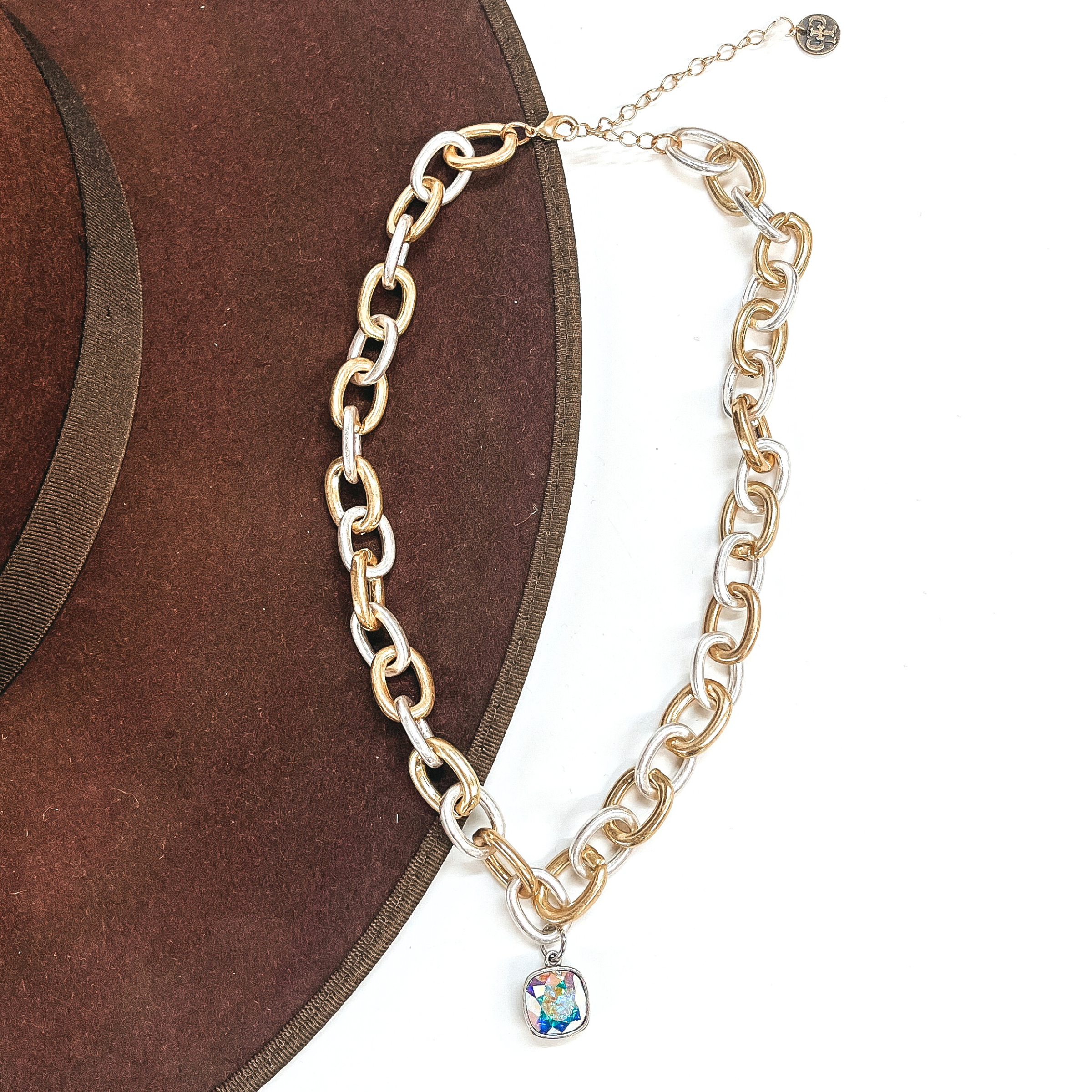 This is silver and gold tone thick linked chain necklace with an AB cushion cut crystal  charm in a silver setting. This necklace is taken laying on a dark brown hat brim  and on a white background.