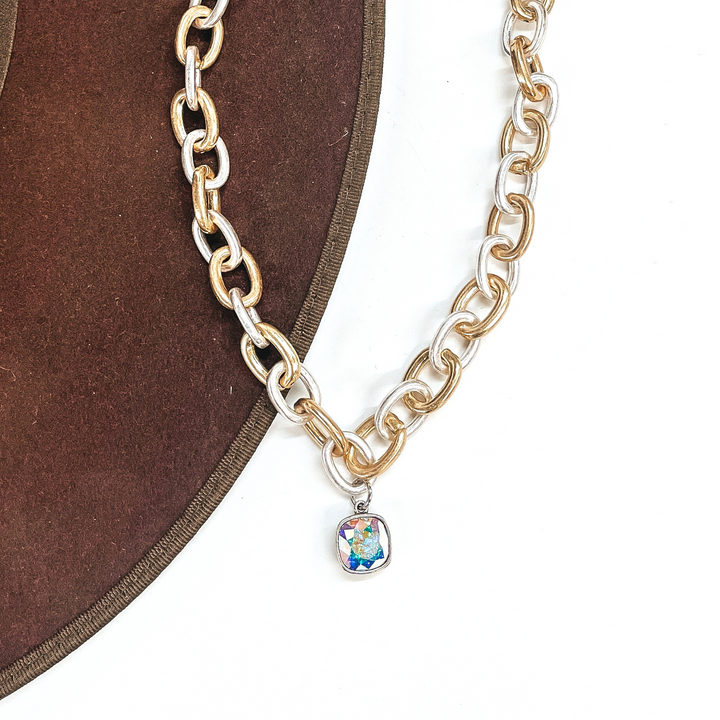 This is silver and gold tone thick linked chain necklace with an AB cushion cut crystal  charm in a silver setting. This necklace is taken laying on a dark brown hat brim  and on a white background.