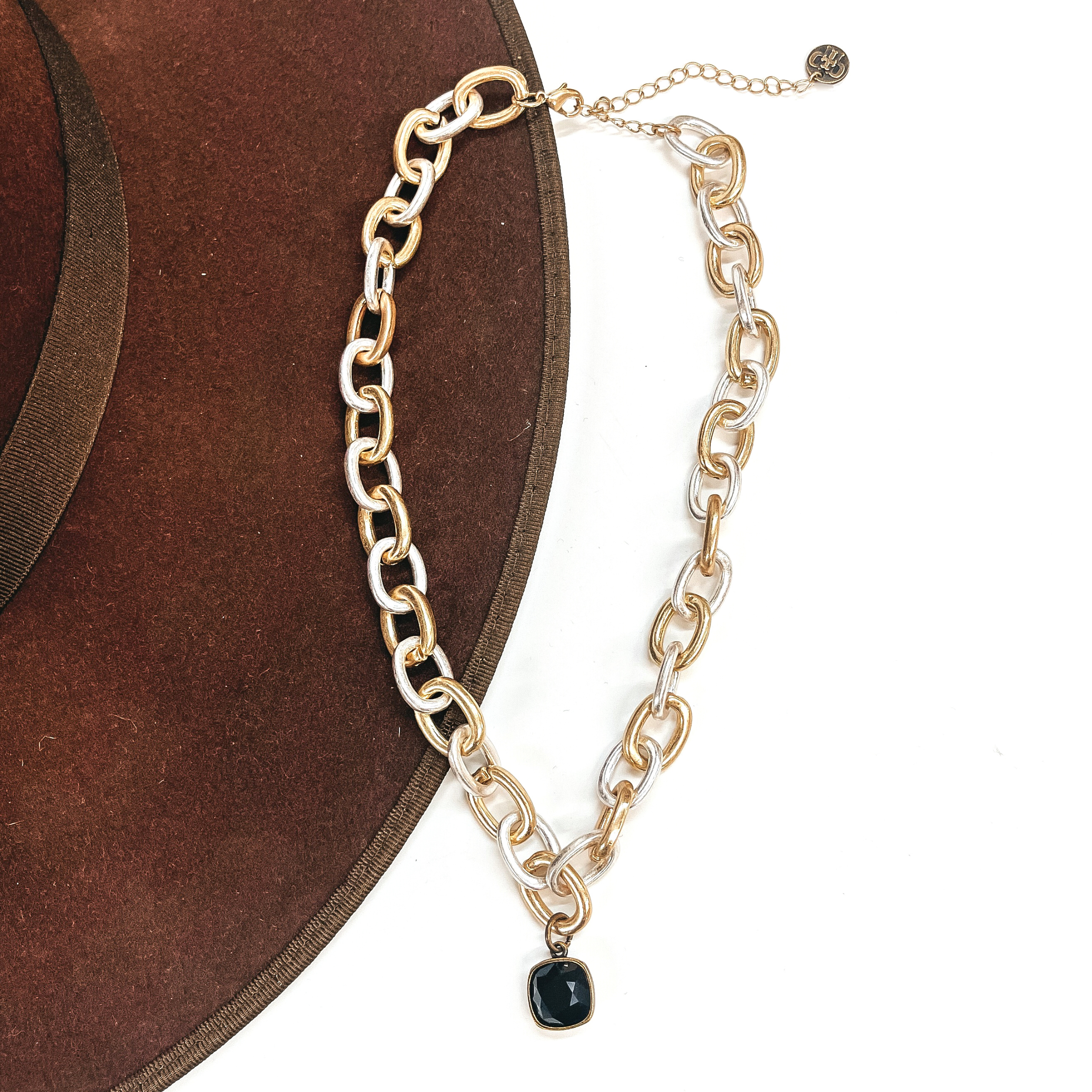 This is silver and gold tone thick linked chain necklace with a black cushion cut crystal  charm in a bronze setting. This necklace is taken laying on a dark brown hat brim  and on a white background.