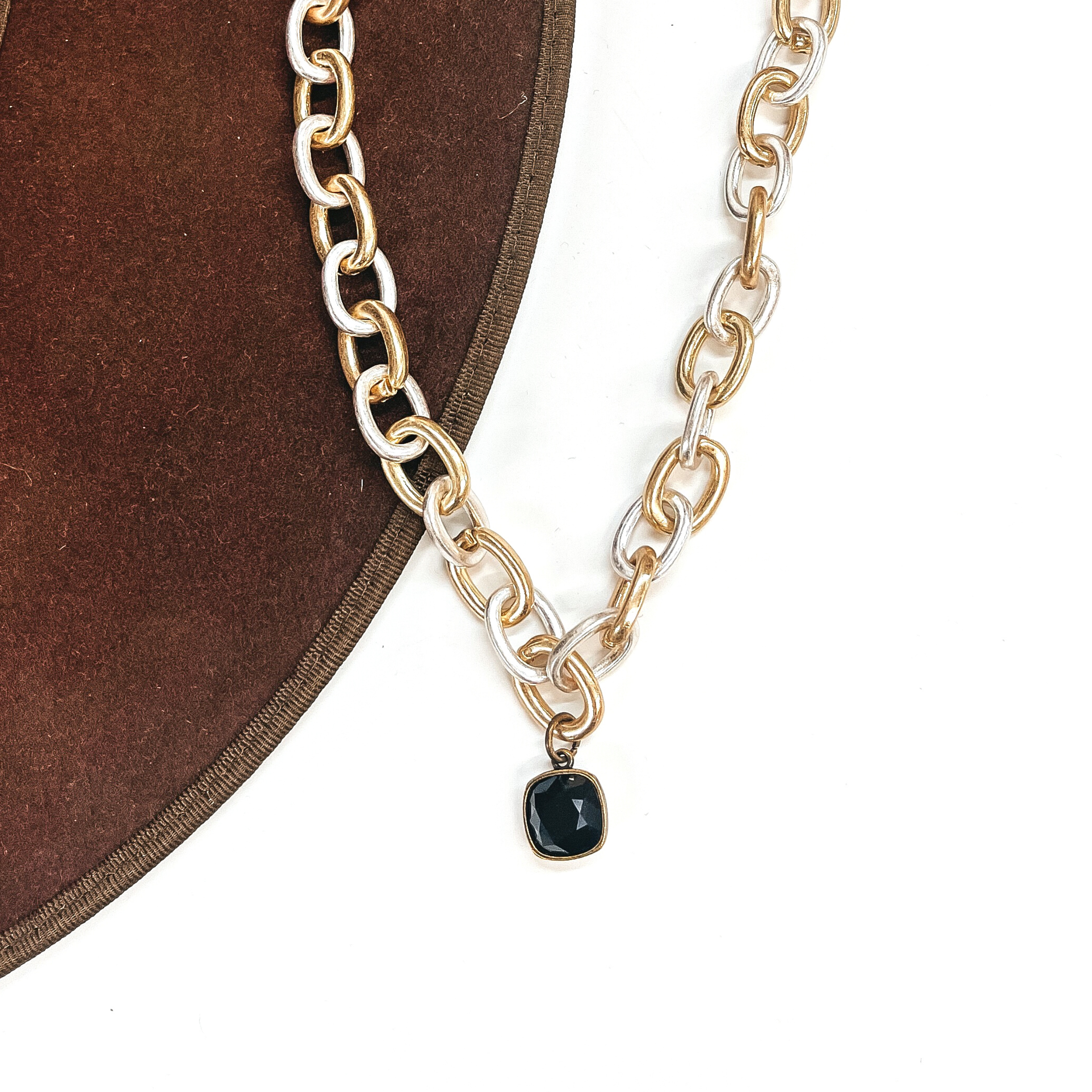 This is silver and gold tone thick linked chain necklace with a black cushion cut crystal  charm in a bronze setting. This necklace is taken laying on a dark brown hat brim  and on a white background.