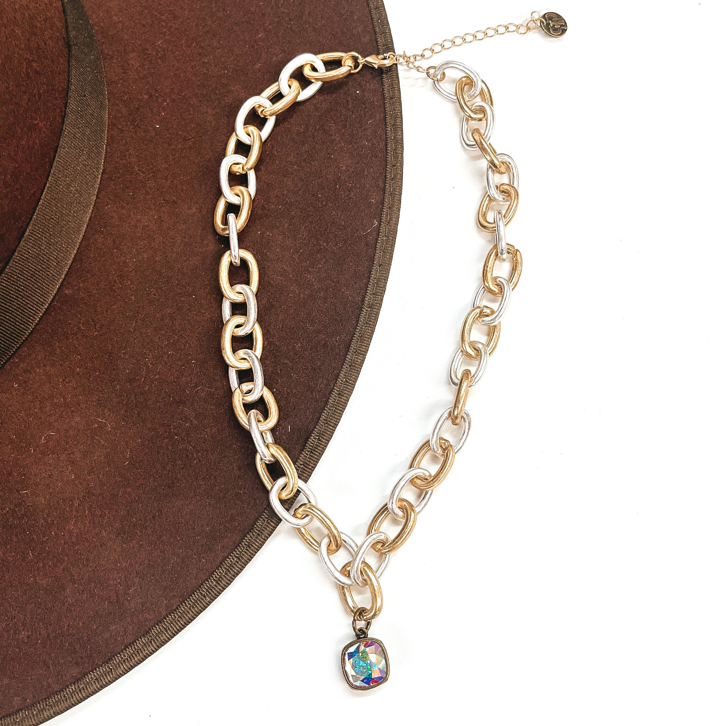 This is silver and gold tone thick linked chain necklace with an AB cushion cut crystal  charm in a bronze setting. This necklace is taken laying on a dark brown hat brim  and on a white background.