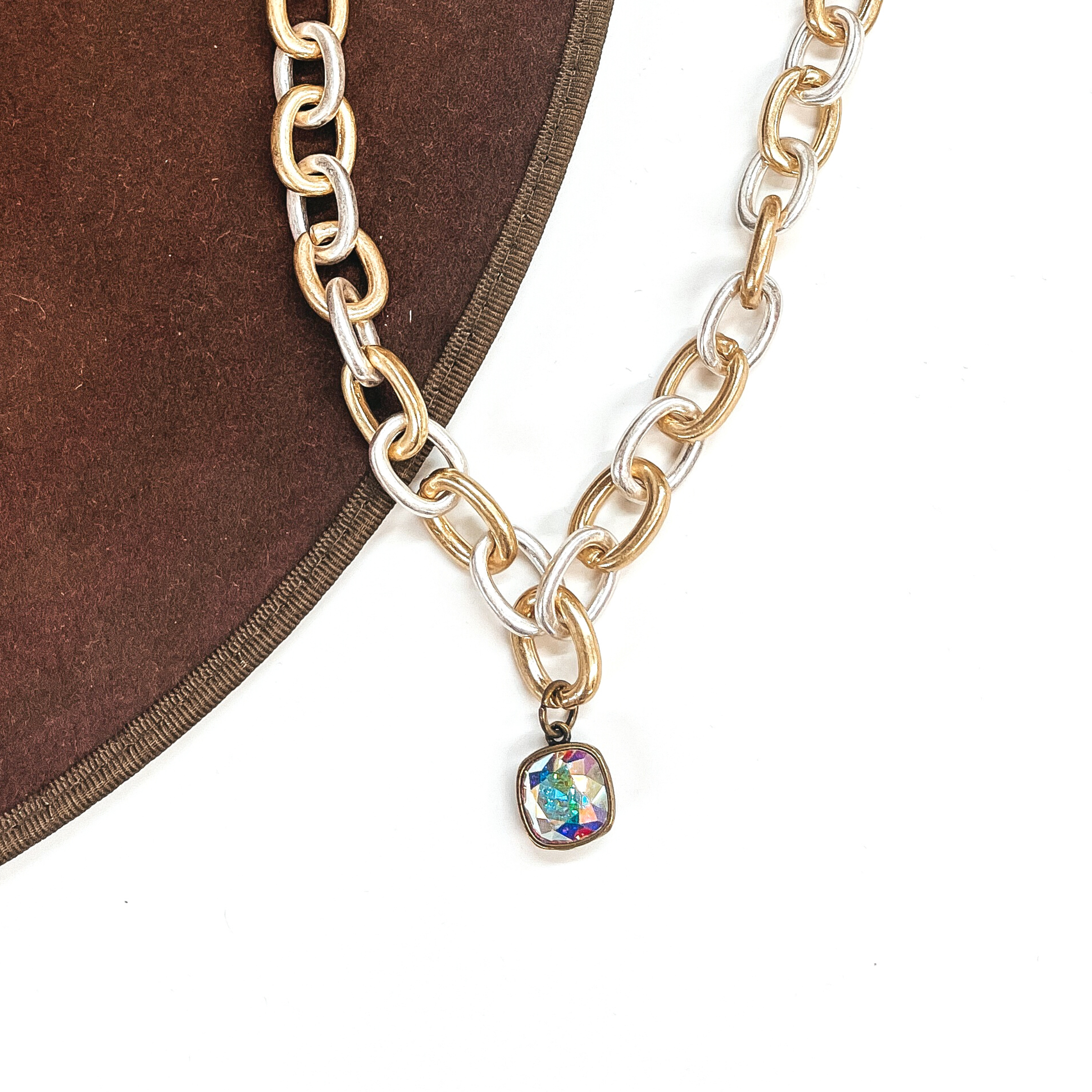 This is silver and gold tone thick linked chain necklace with an AB cushion cut crystal  charm in a bronze setting. This necklace is taken laying on a dark brown hat brim  and on a white background.