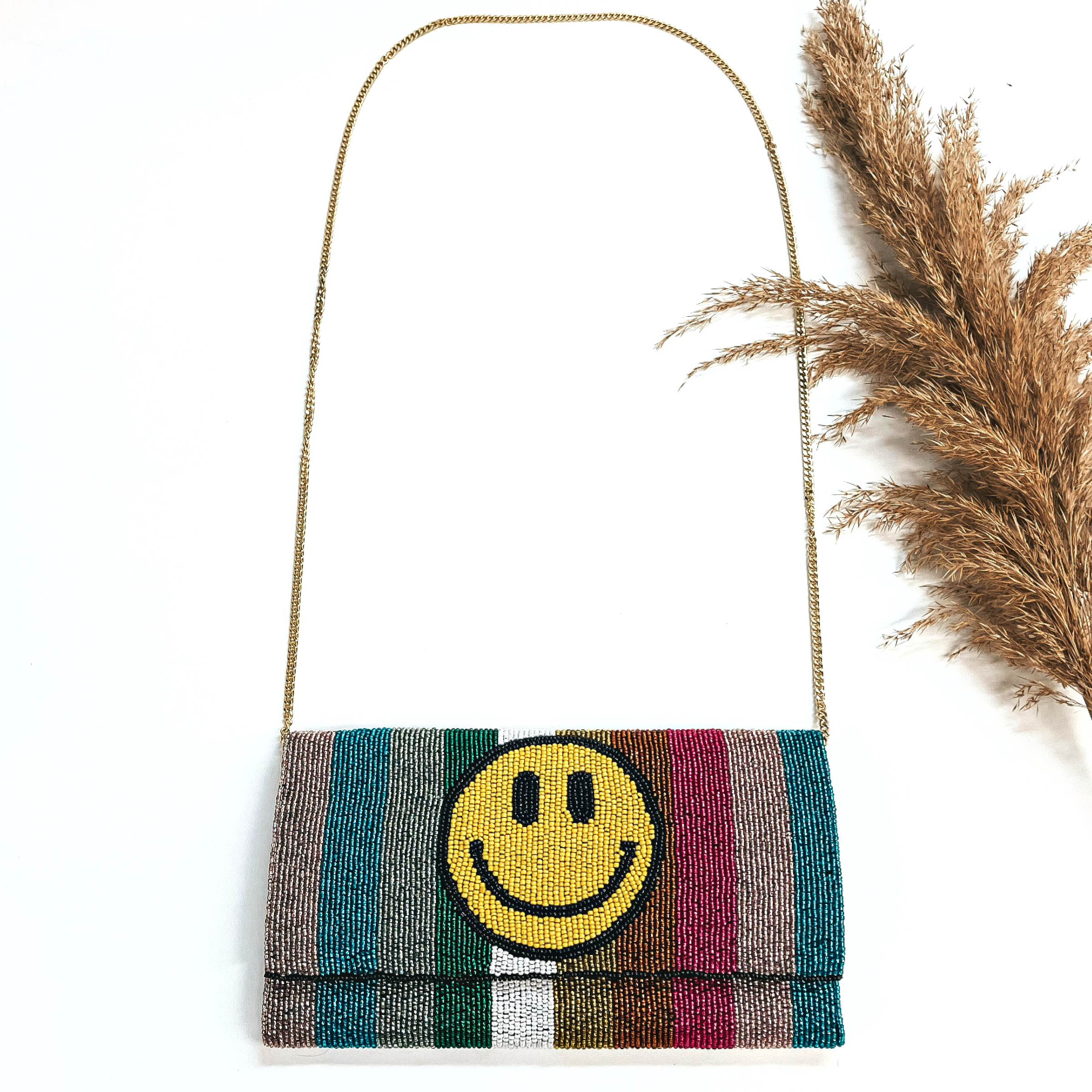 This is a seedbeaded clutch purse with a gold tone chain. This bag is multicolor with  lines and a yellow happy face. Multicolor lines in teal, rose gold, fuchsia, copper,  yellow, white, green, and mint. This bag  is taken laying on a white background with a brown plant in the side as decor.